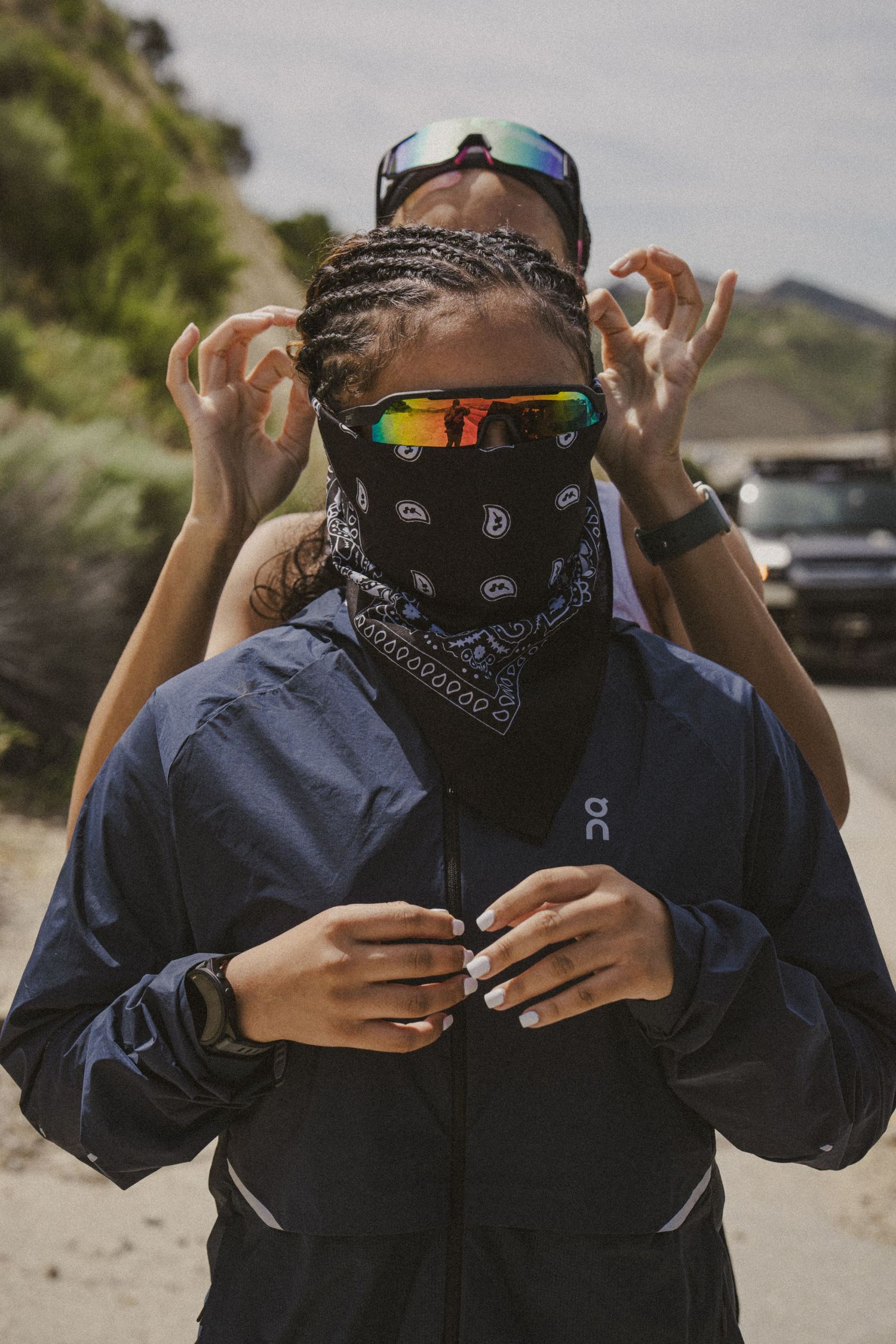 Person wearing bandana face mask and On Running jacket with colorful sunglasses outdoors on a dirt path.