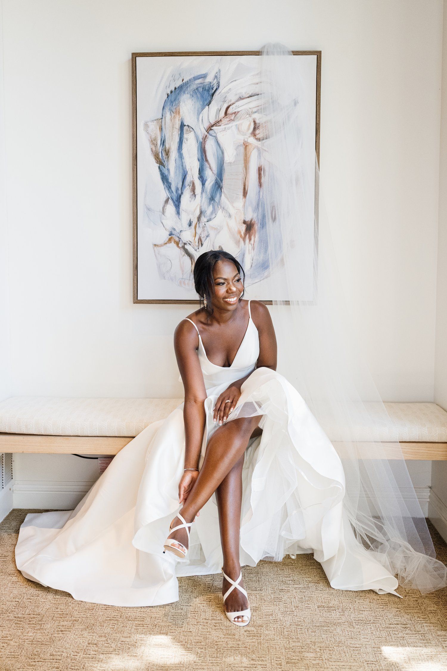 A bride in a white gown adjusts her shoe while sitting beneath an abstract blue watercolor painting.