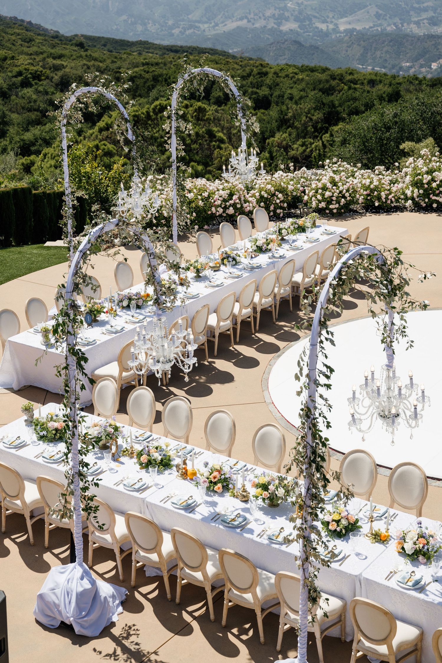 Elegant outdoor wedding reception with long white tables, floral arches, chandeliers, and mountain views.