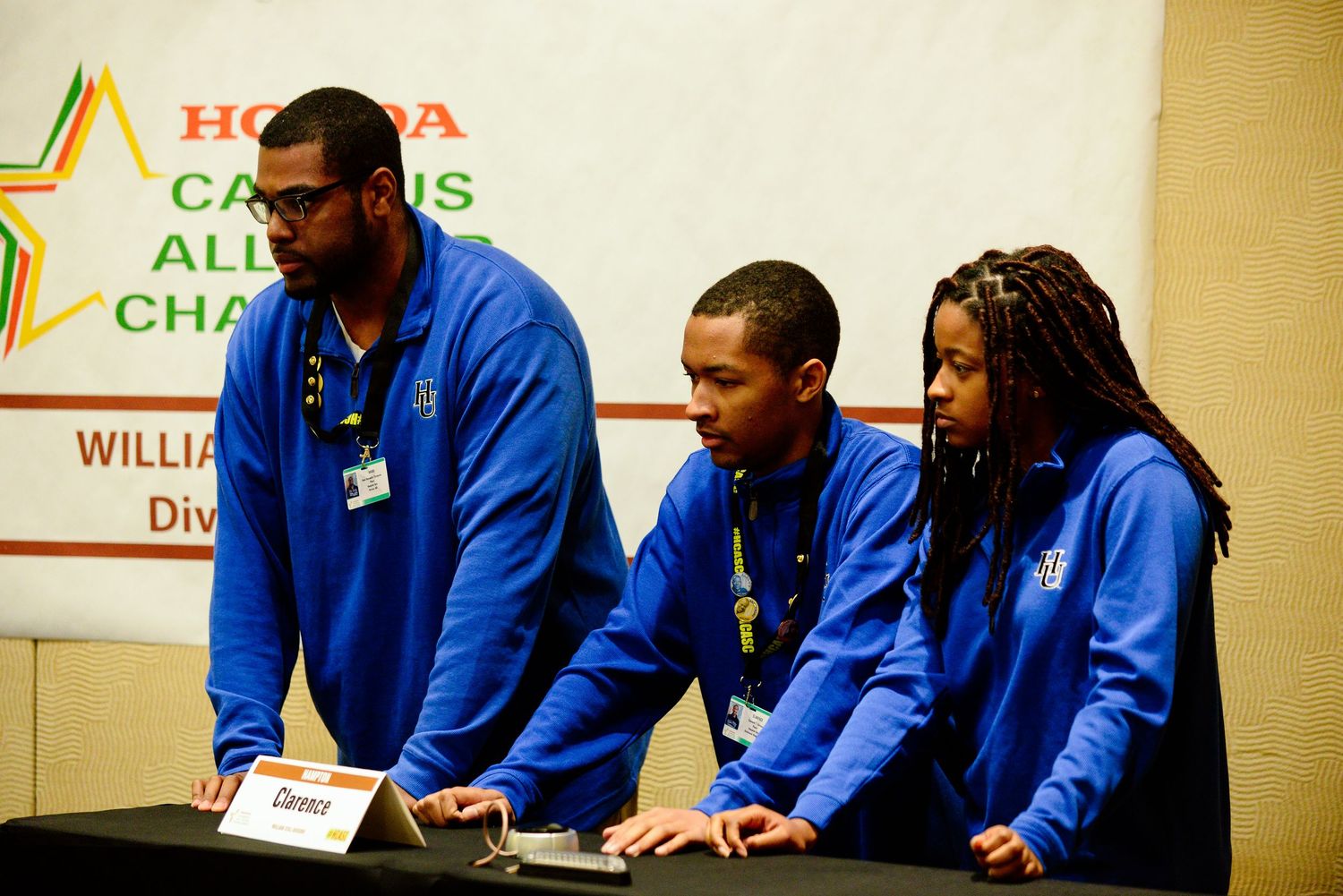 Row of students in blue uniforms seated at table during academic presentation or competition.