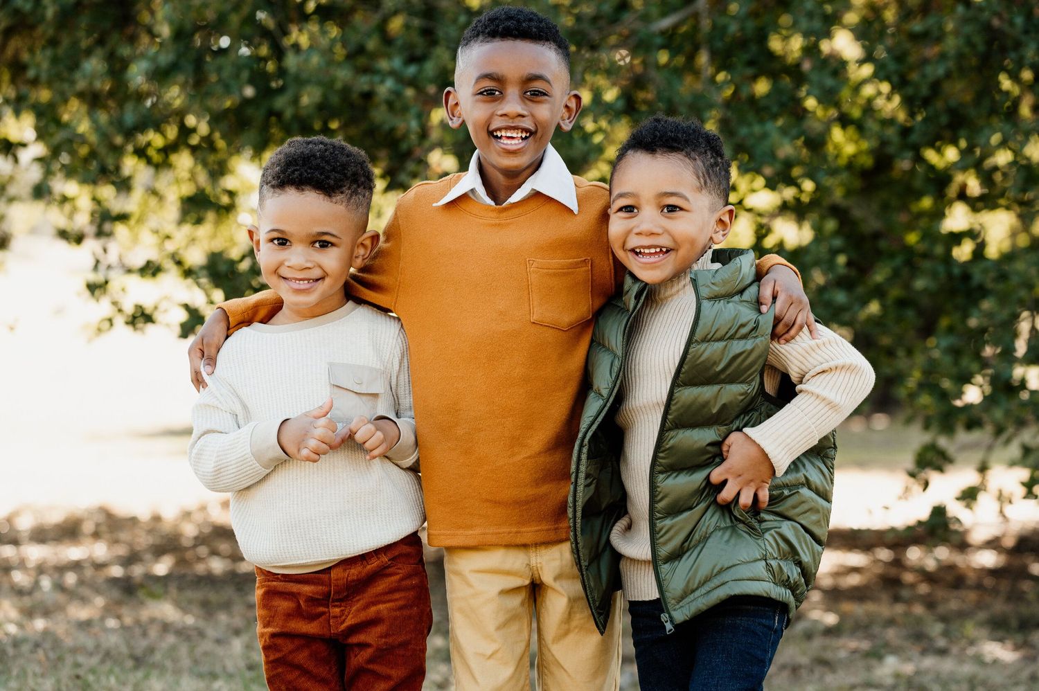 Three young boys in casual fall clothing smile together outdoors.