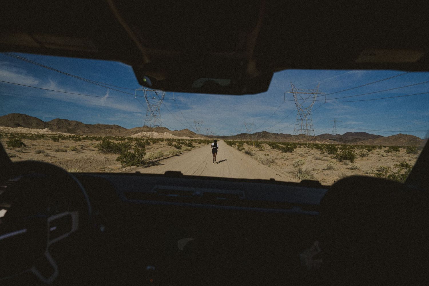 View from inside a vehicle of a lone person walking on a desert road with power lines and mountains under a blue sky.