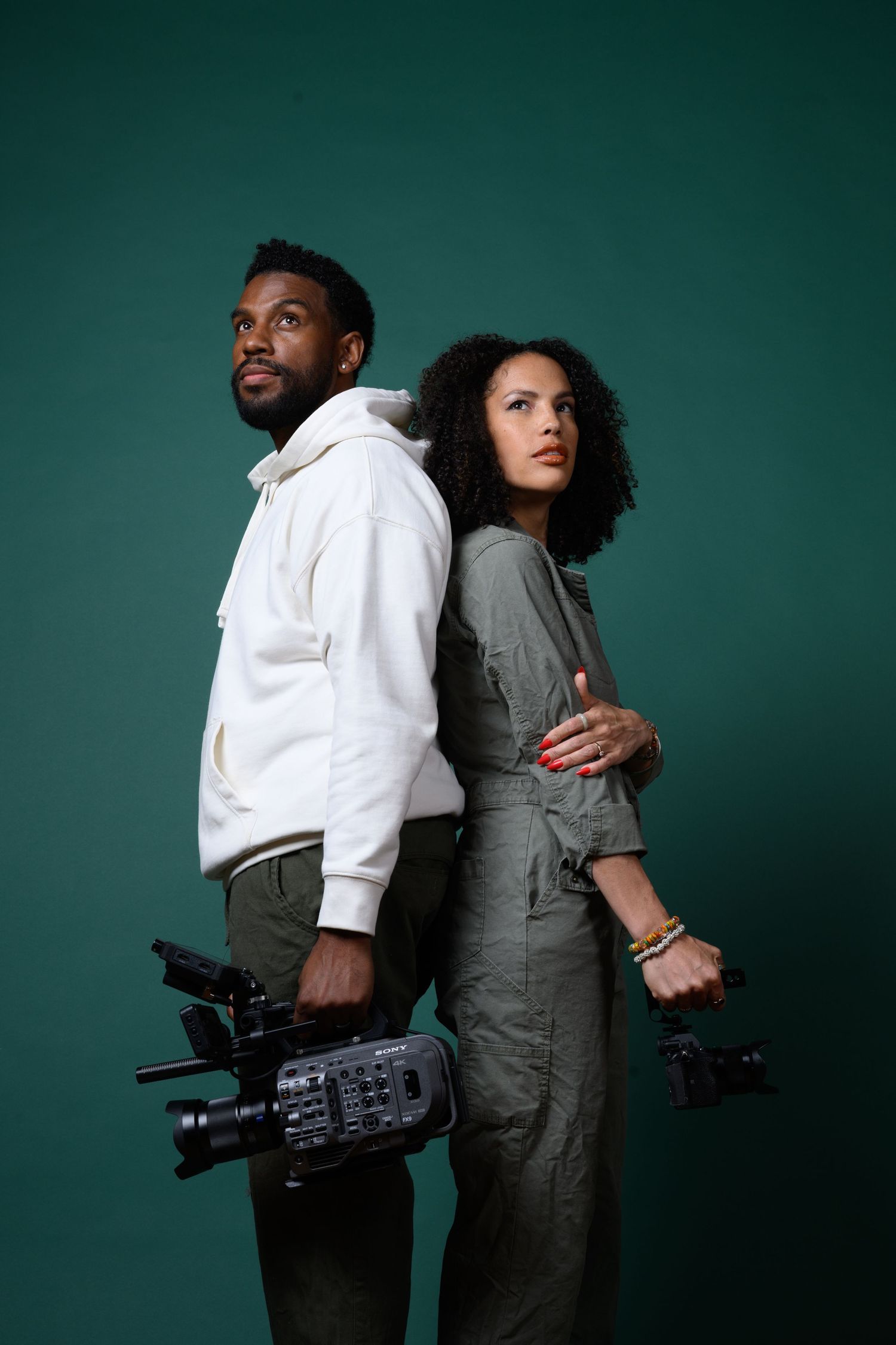 Two photographers stand back-to-back holding professional video cameras against a dark green studio background.