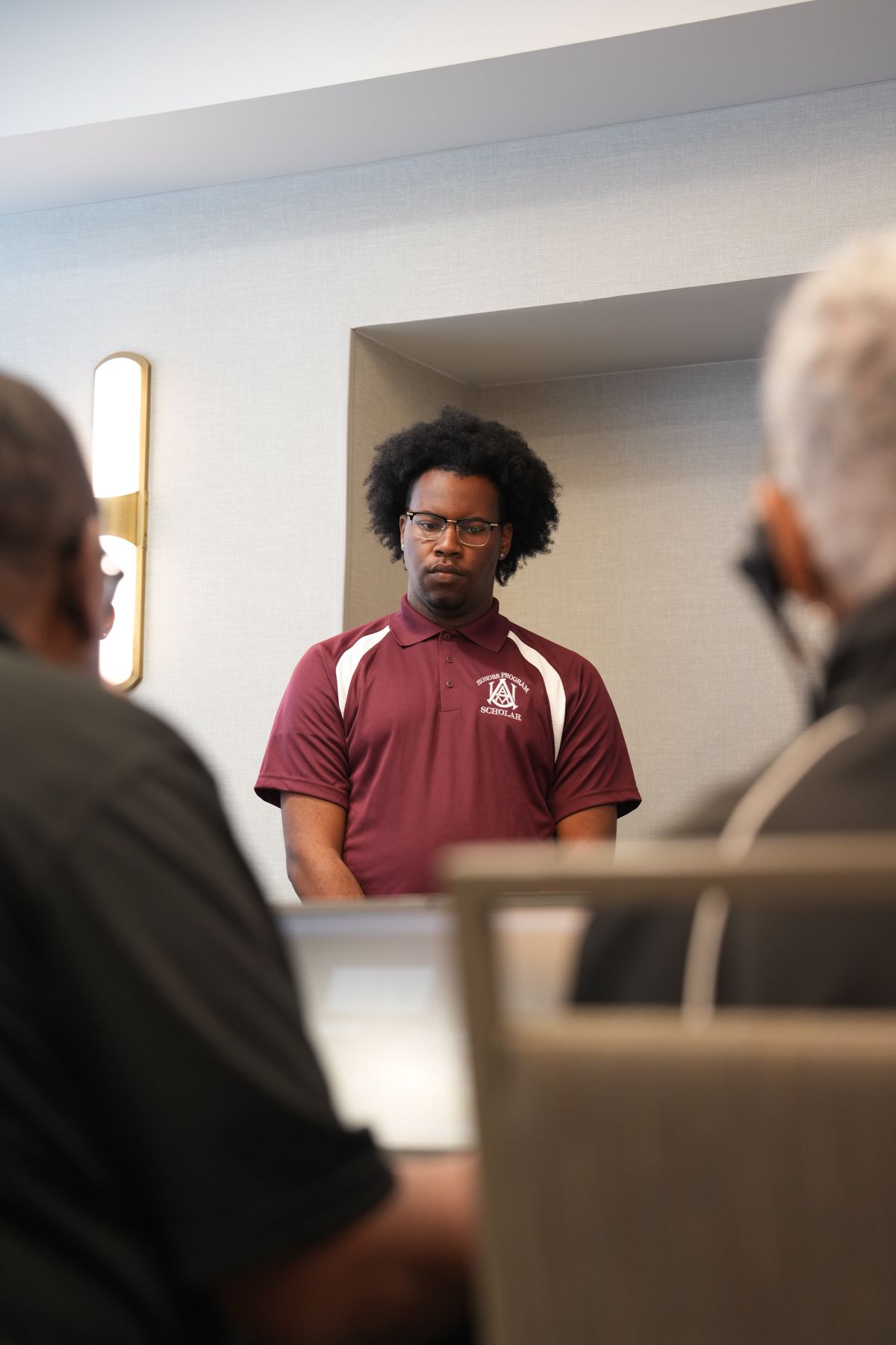 Young man in maroon polo shirt with glasses stands attentively in modern interior setting during a meeting.
