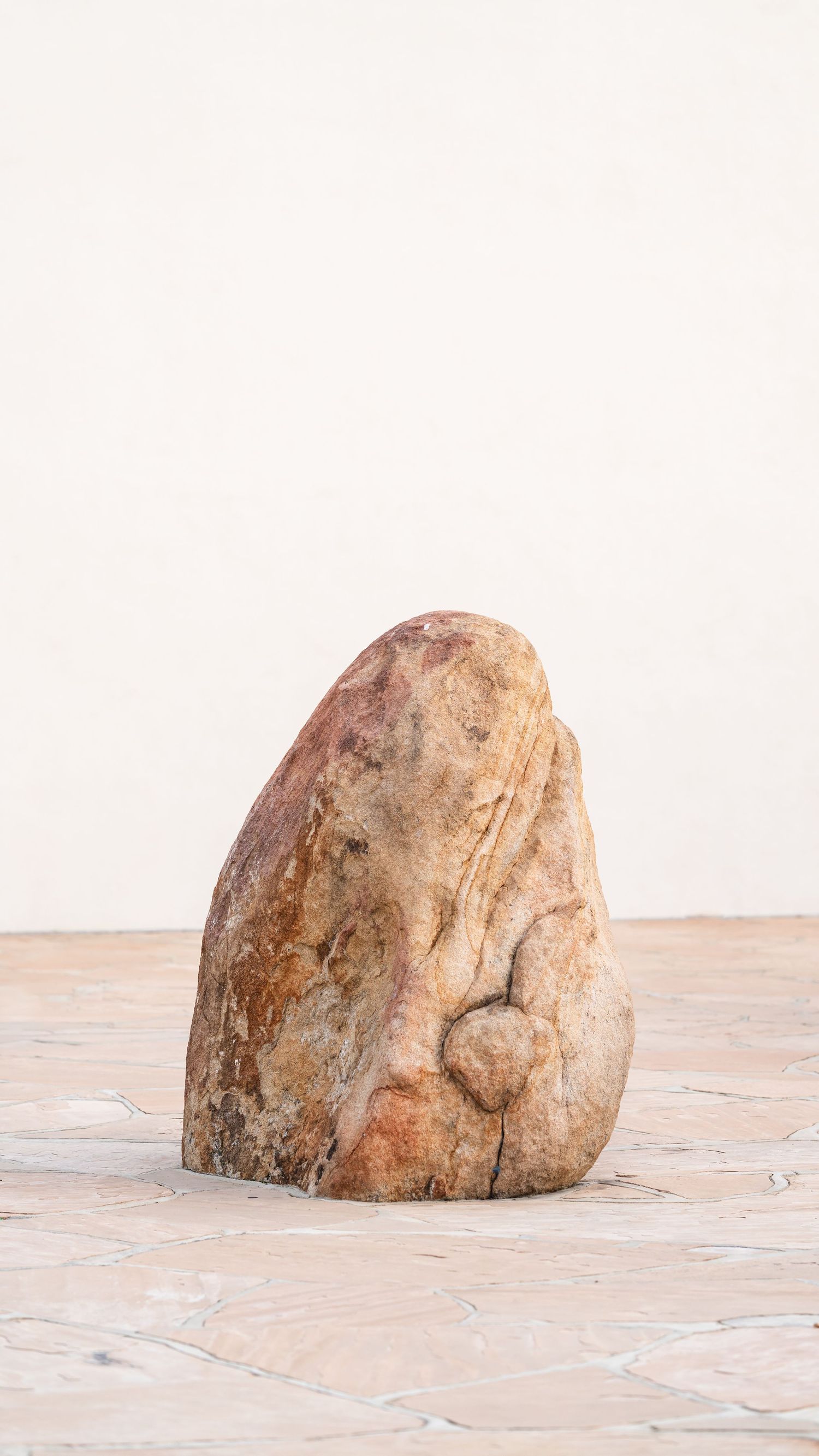 A weathered sandstone rock sculpture with organic form sits on a tiled floor against a white wall.