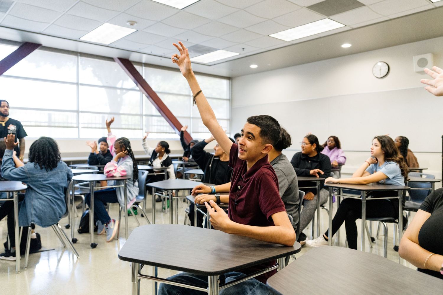 Students raising their hands in a classroom with fluorescent lighting and white tables.