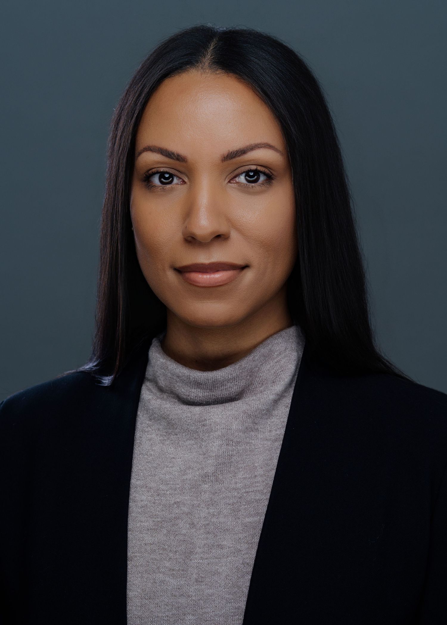 Professional headshot of woman in black blazer and grey turtleneck against dark background.