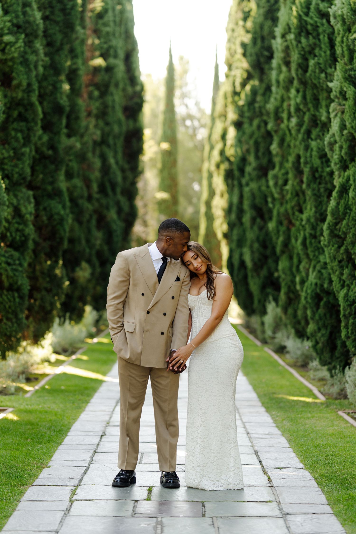 Newlyweds share intimate moment on cypress-lined path in formal attire.