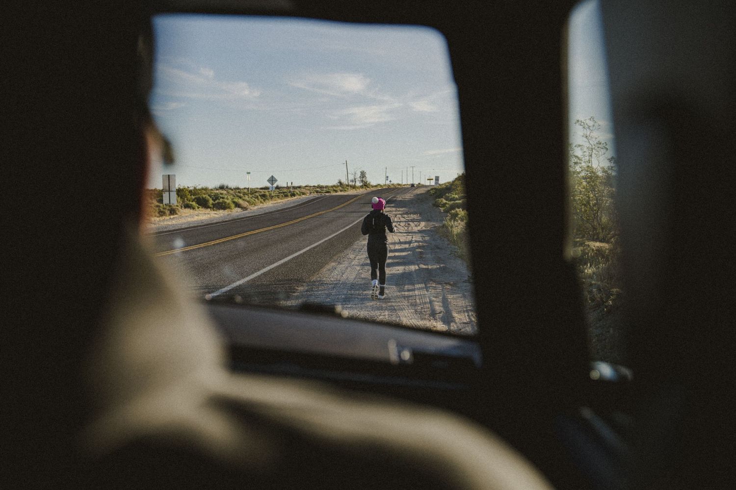 Person running on a desert highway viewed through a car rear window on a sunny day.