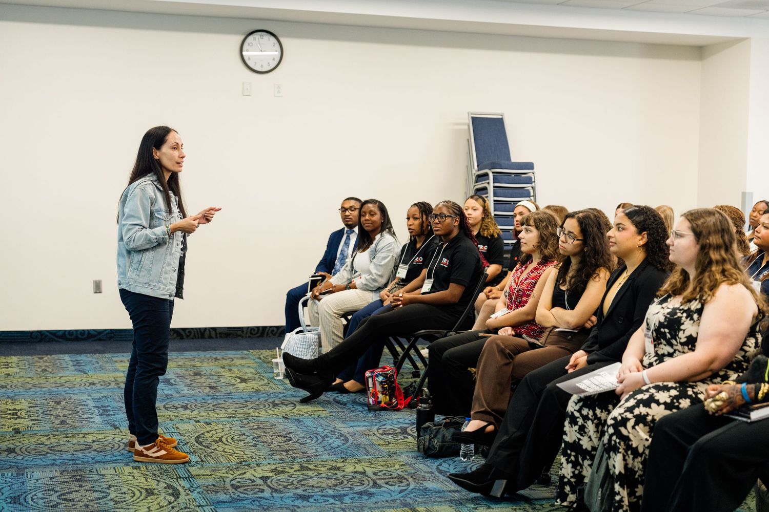 A woman presents to a diverse group of students seated in rows during an indoor educational seminar or workshop.