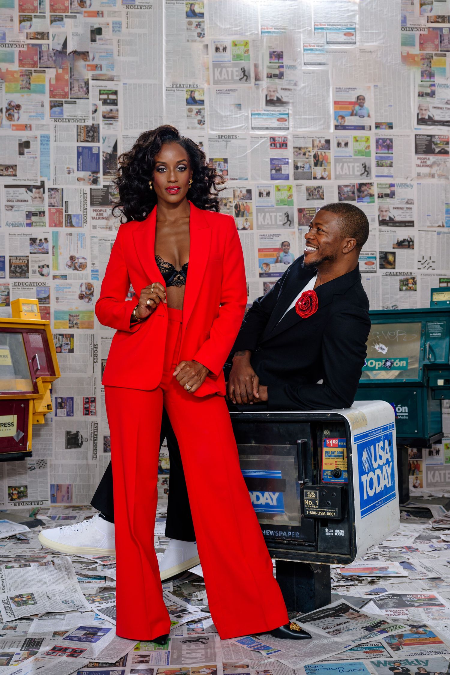 Fashion photoshoot featuring bold red suit against a wall covered in newspaper clippings.