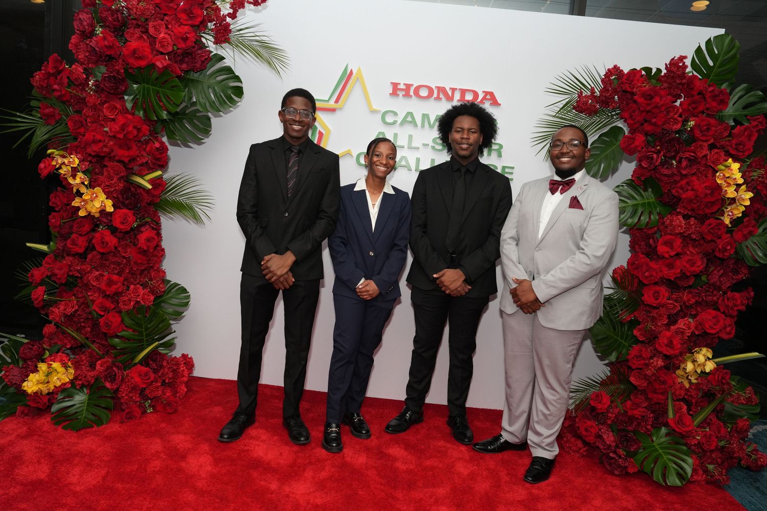 Four men posing on a red carpet at the Honda Campus All-Star Challenge, surrounded by red floral decorations.