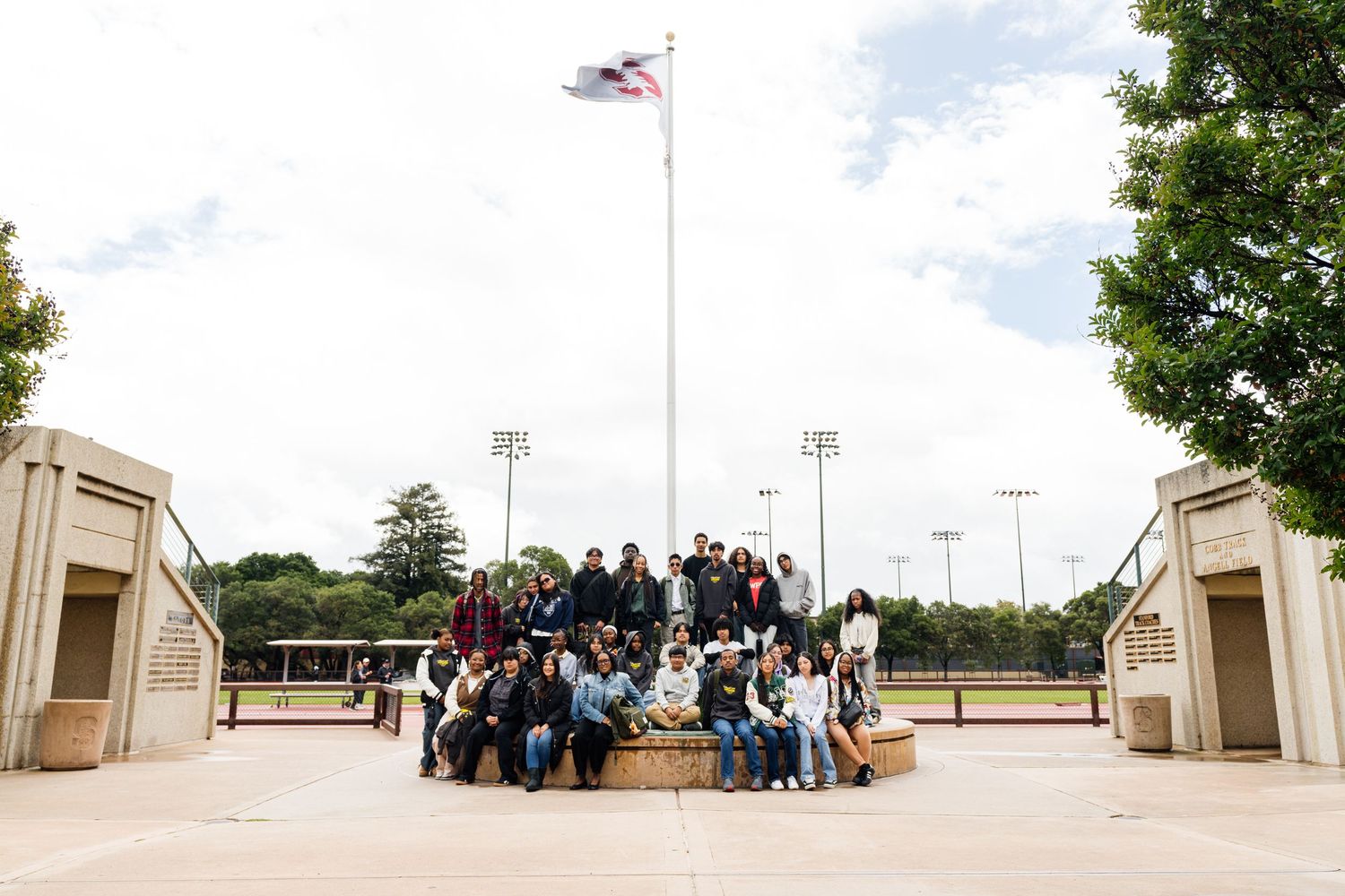 Large group of students posing together outdoors near a flagpole at a sports complex on a cloudy day.