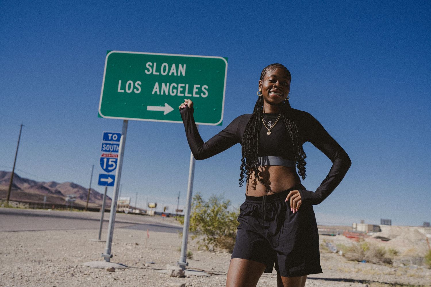 A woman poses confidently beside a green highway sign pointing to Los Angeles in a sunny desert landscape.