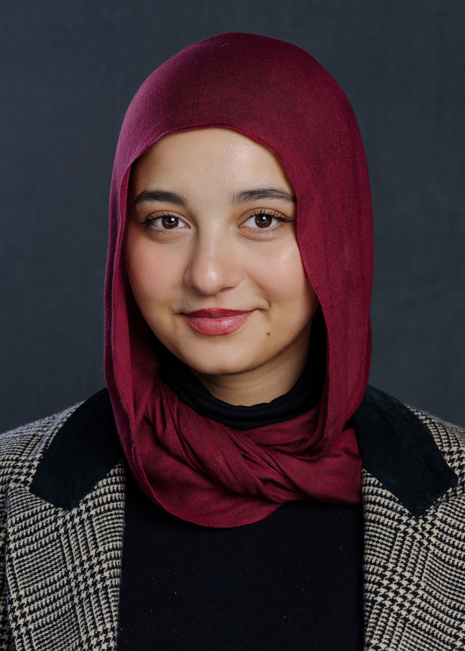 Professional portrait of someone wearing a burgundy hijab and plaid blazer against a dark background.