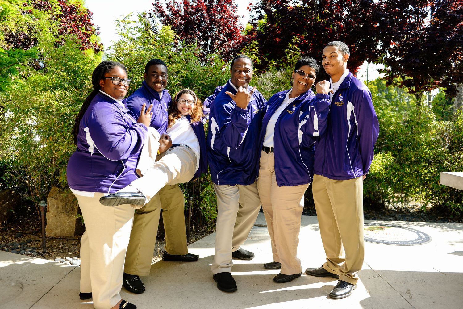 Group of students in khaki pants and navy shirts sitting outdoors on steps near flowering trees.
