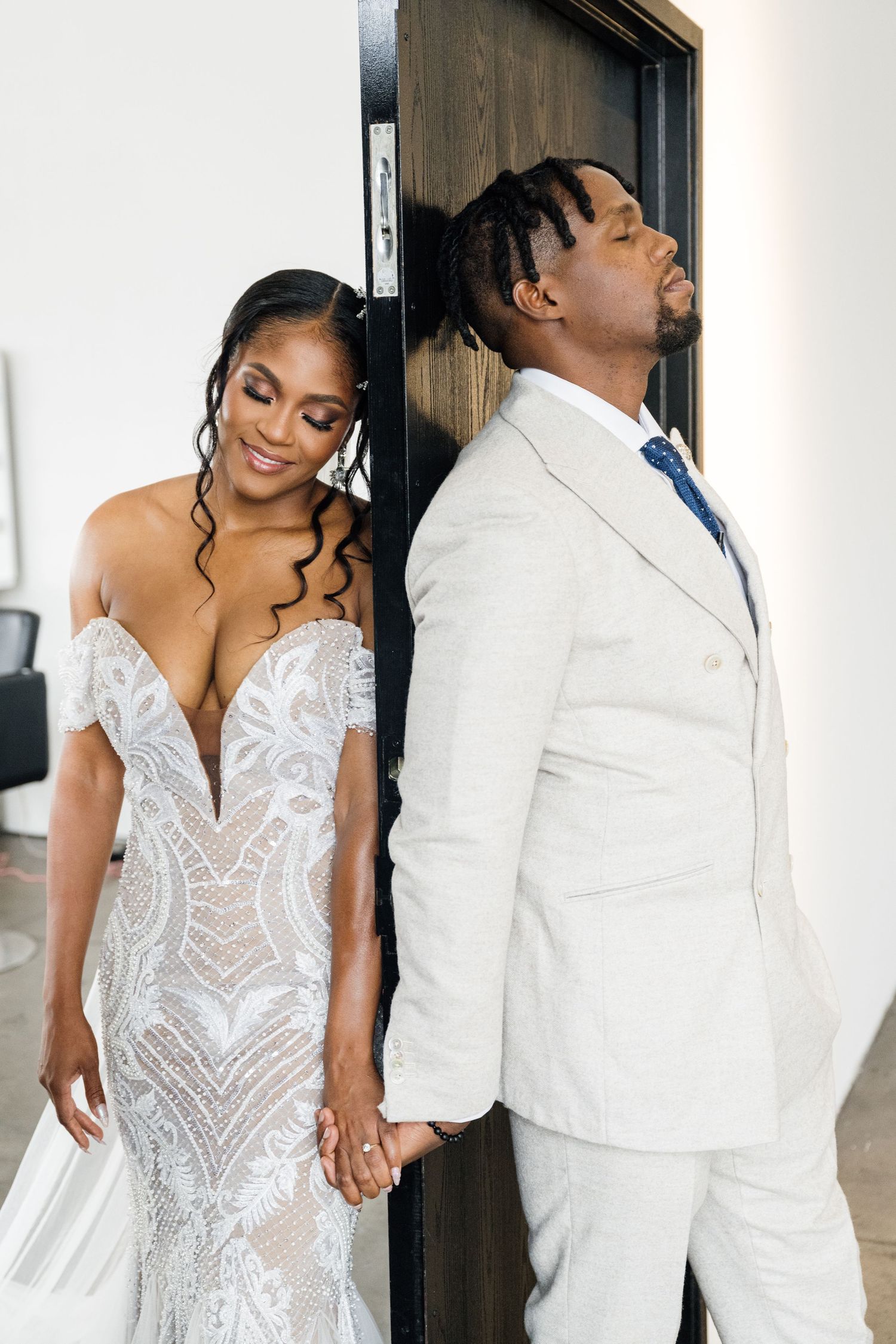 Bride in white lace gown poses with partner in white suit against neutral backdrop.