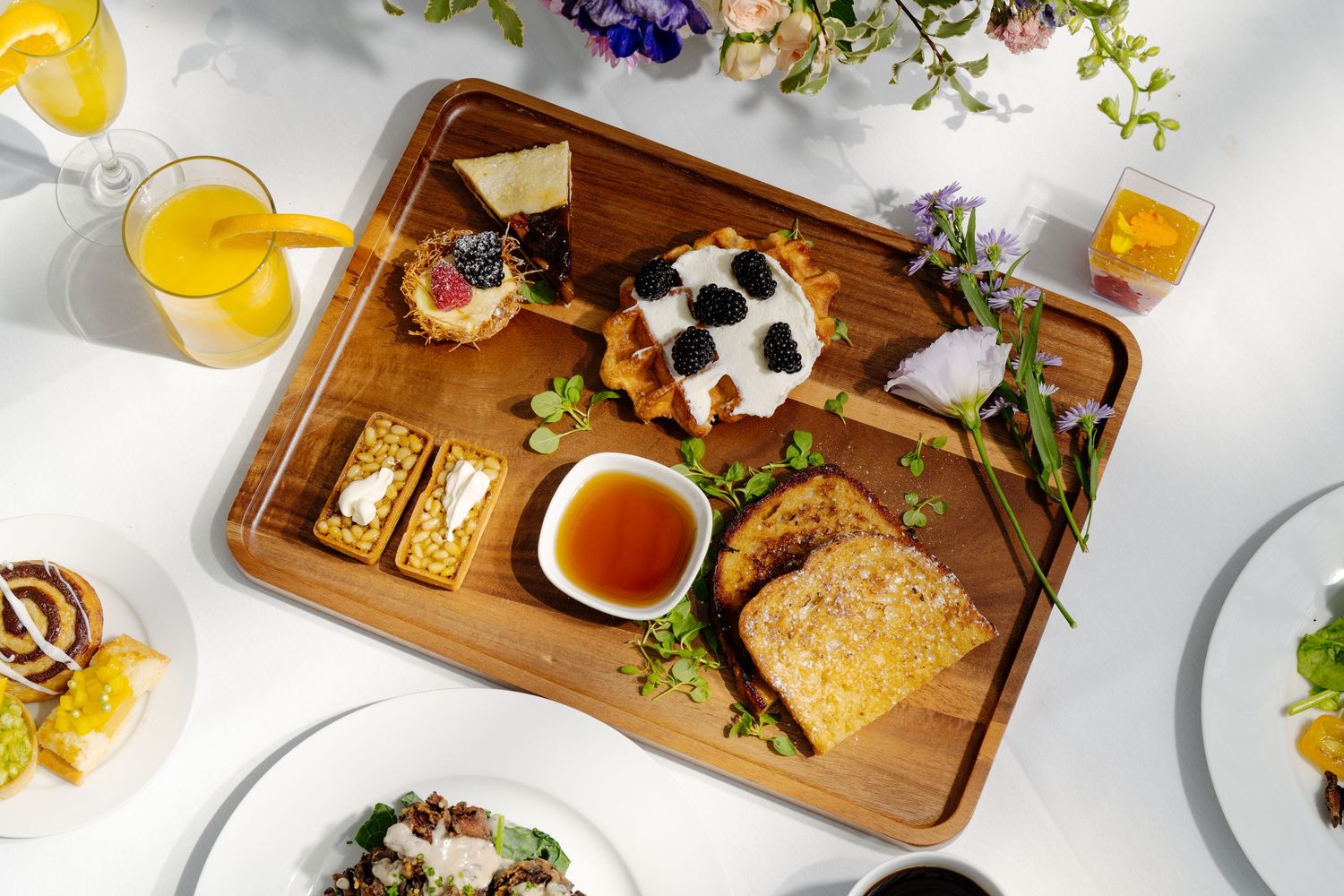 Elegant brunch board with waffles, blackberries, honey, toast, and crackers on a wooden tray with fresh flowers.