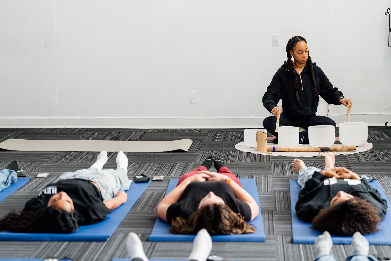 Instructor leads a sound bath session with singing bowls while participants lie on blue yoga mats in a studio.