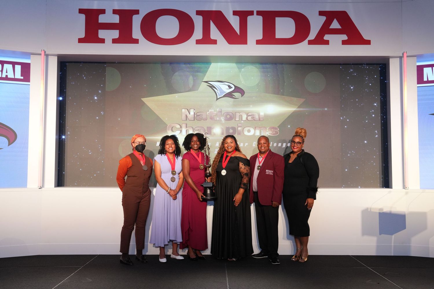 Group of women posing on stage at Honda National Awards ceremony holding trophy under spotlight.