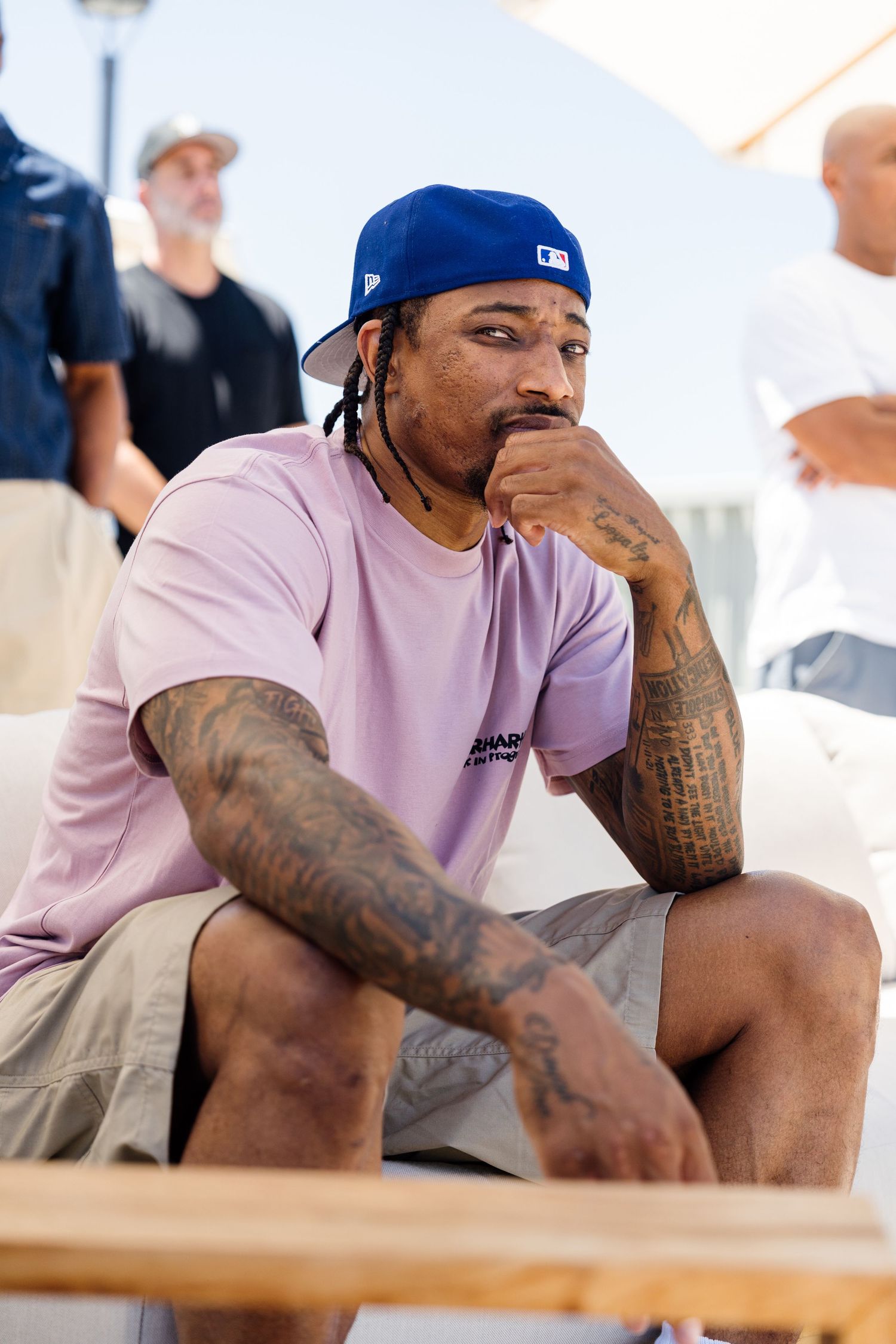 Tattooed man in blue cap and pink tee sits pensively outdoors, hand on chin, with people in background.