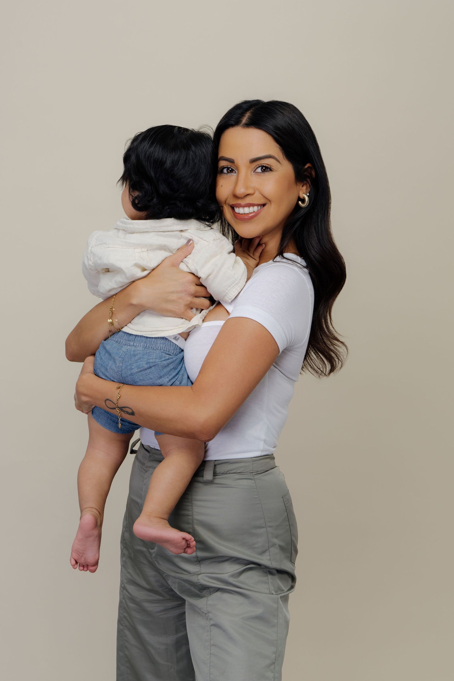 A parent in gray overalls and white shirt holds a baby in white clothing against a neutral background.
