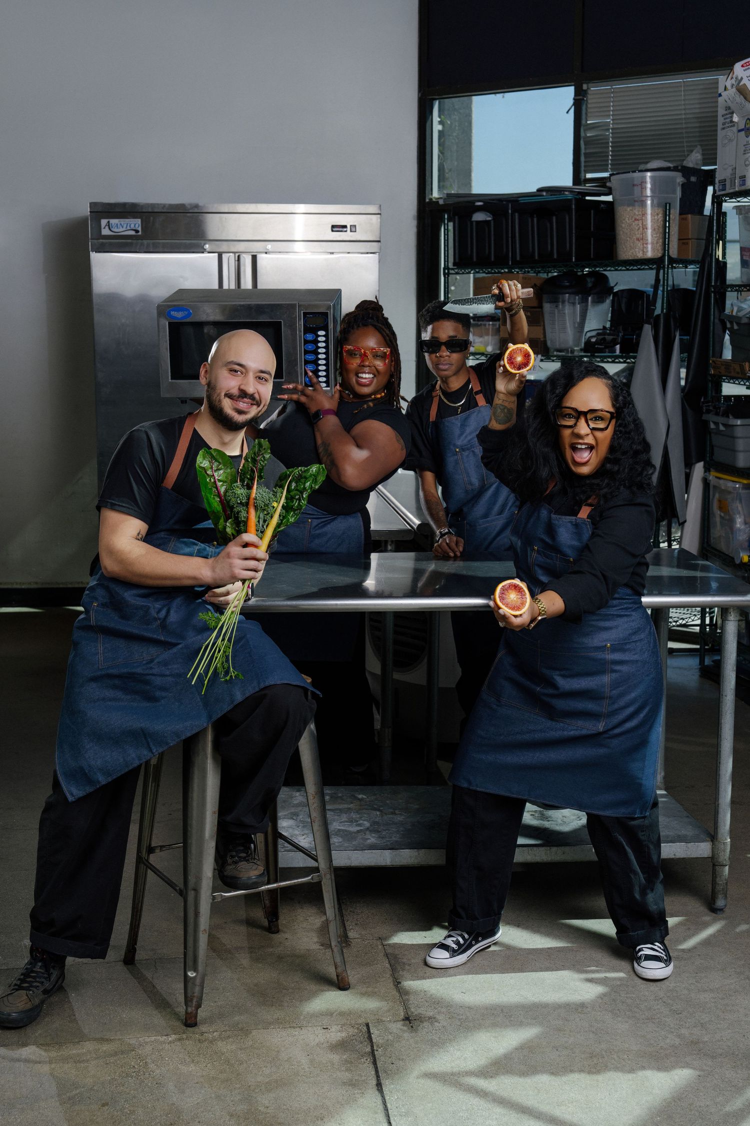 Two chefs in blue uniforms playfully pose with vegetables in a commercial kitchen setting.
