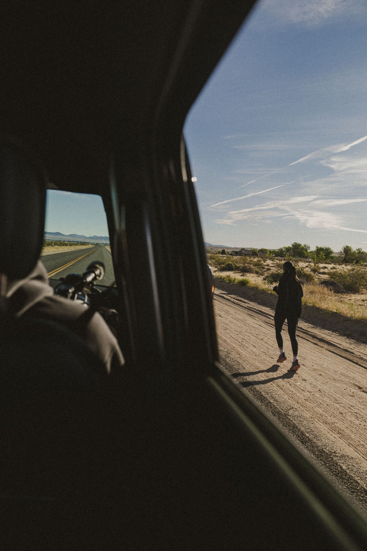 A woman stands on a desert roadside beside a parked vehicle, viewed from inside through the window on a sunny day.