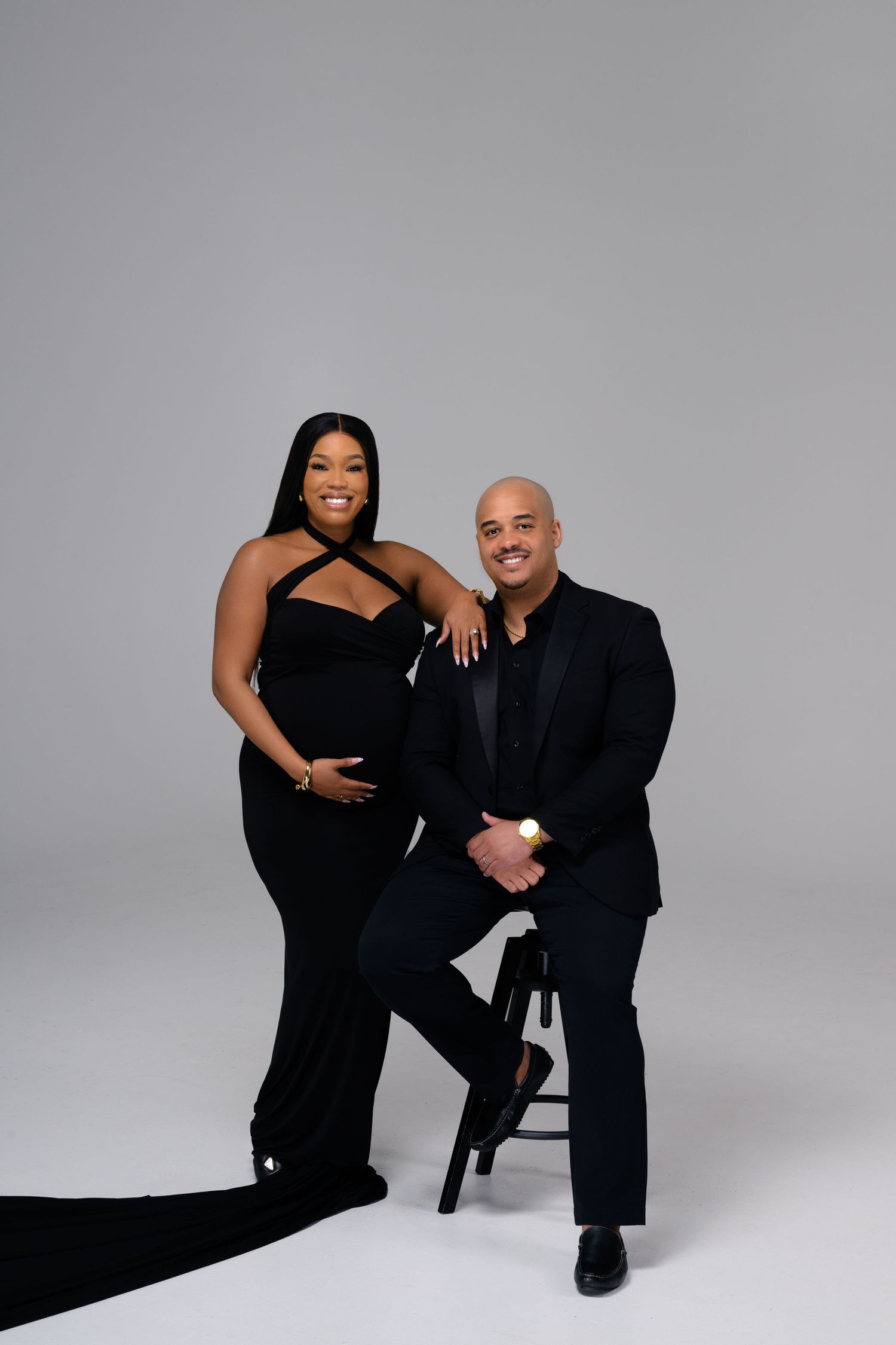 Studio photoshoot featuring couple in matching black outfits against minimalist grey background.