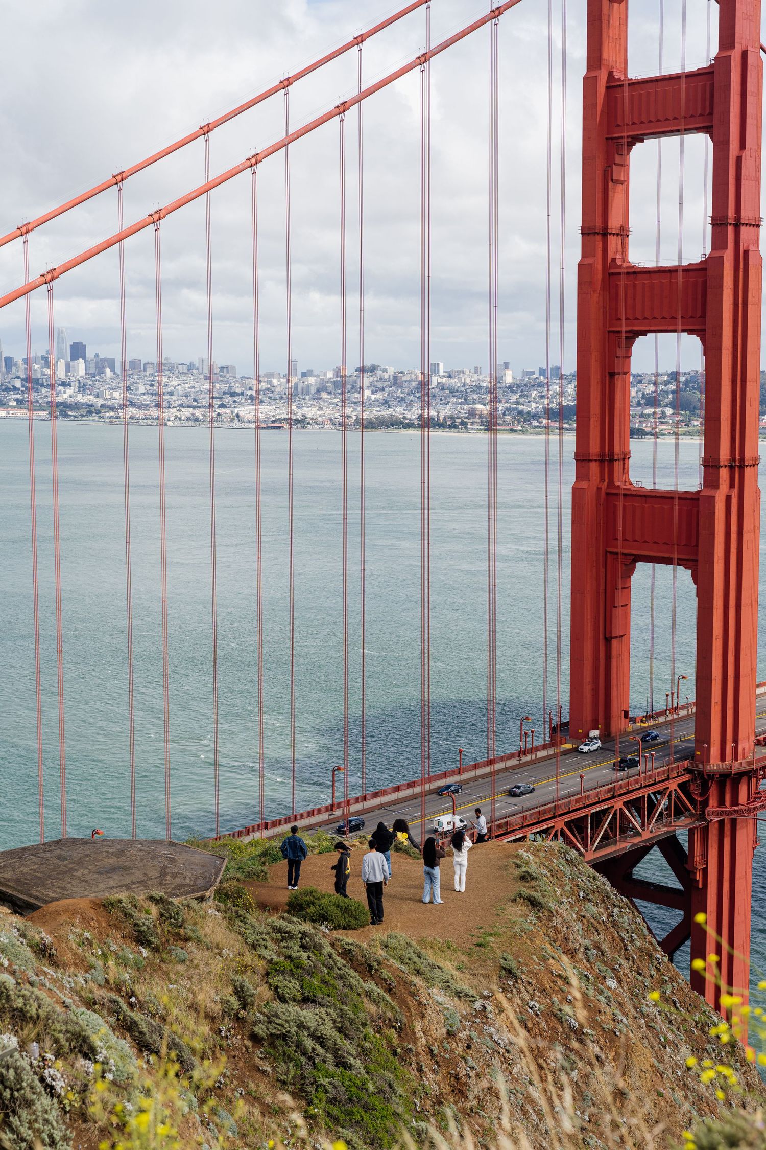 Tourists walk along a path near the iconic red Golden Gate Bridge with San Francisco skyline visible across the bay.