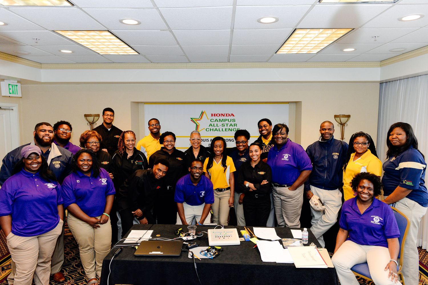 Large group photo of students and faculty in blue uniforms gathered in conference room setting.