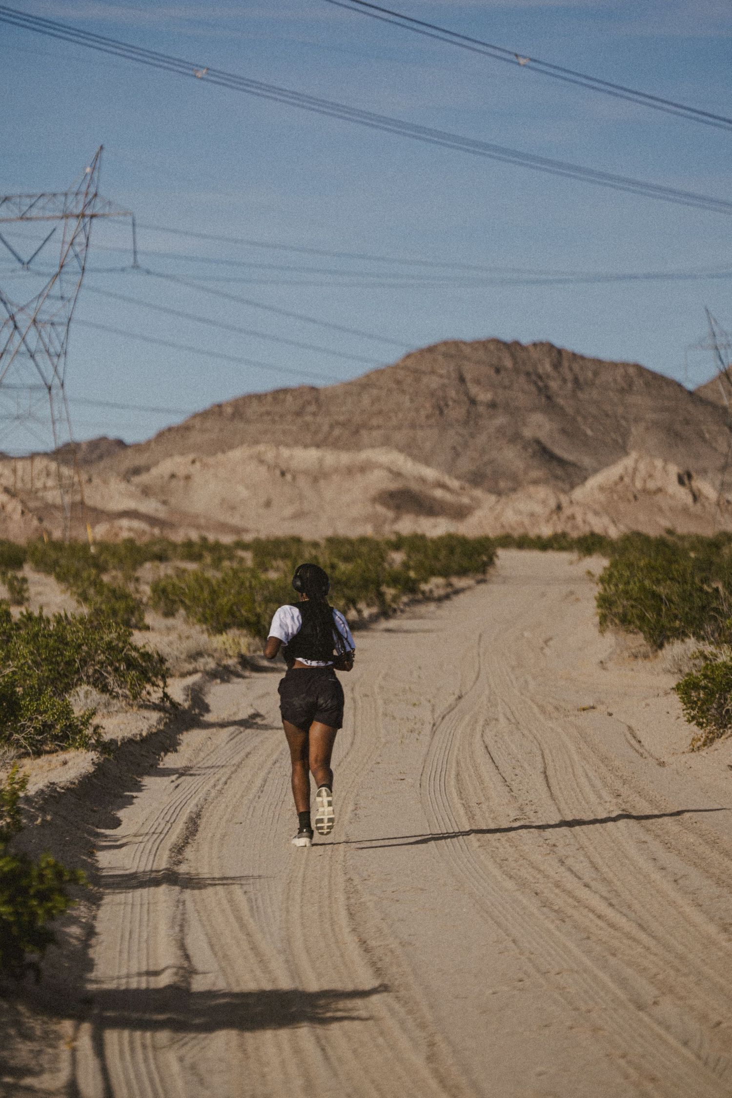 Woman jogging on sandy desert dirt road with power lines and rocky mountains in the background under clear blue sky.