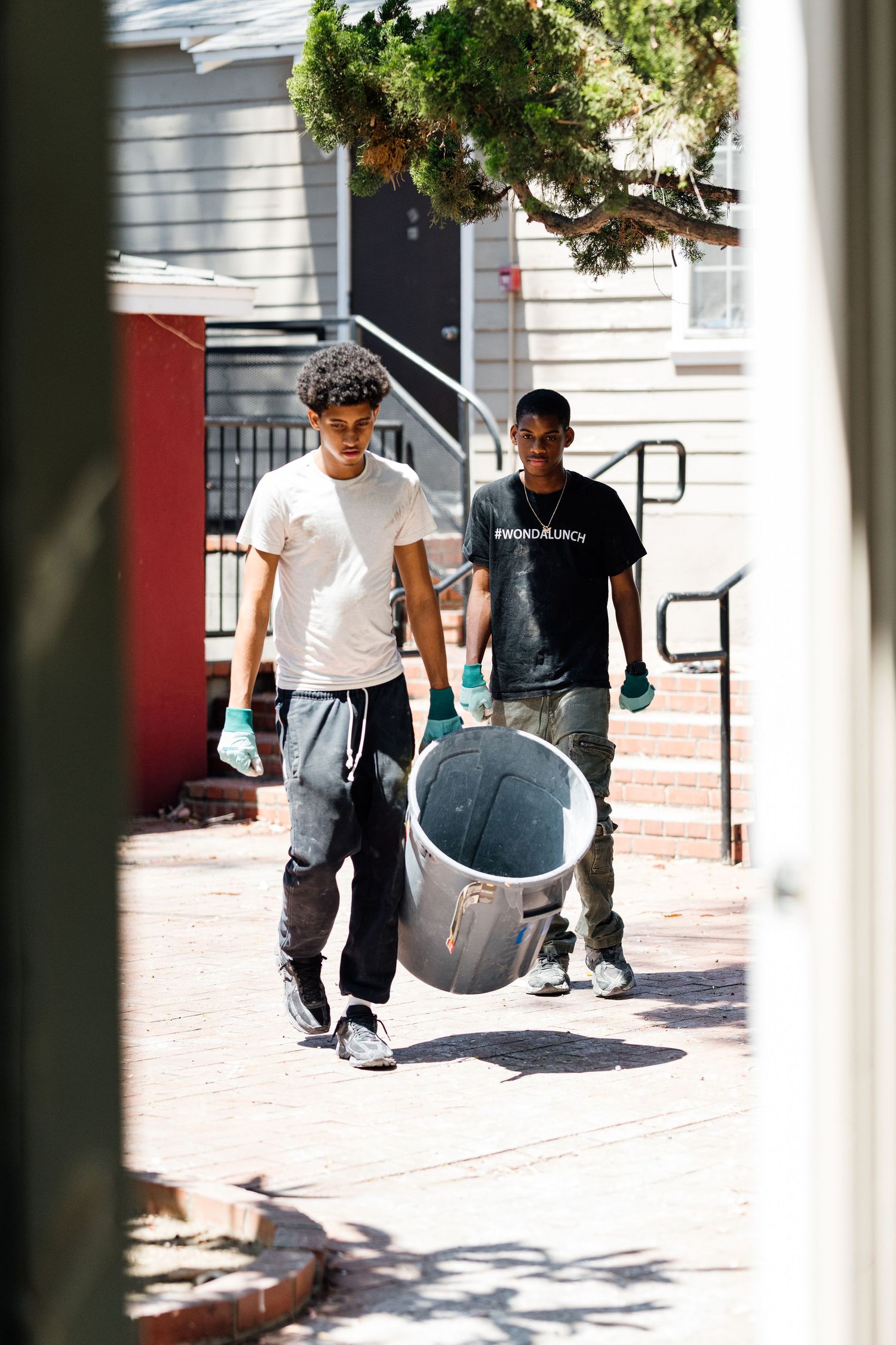 People carrying and moving equipment on a sunny sidewalk near buildings.