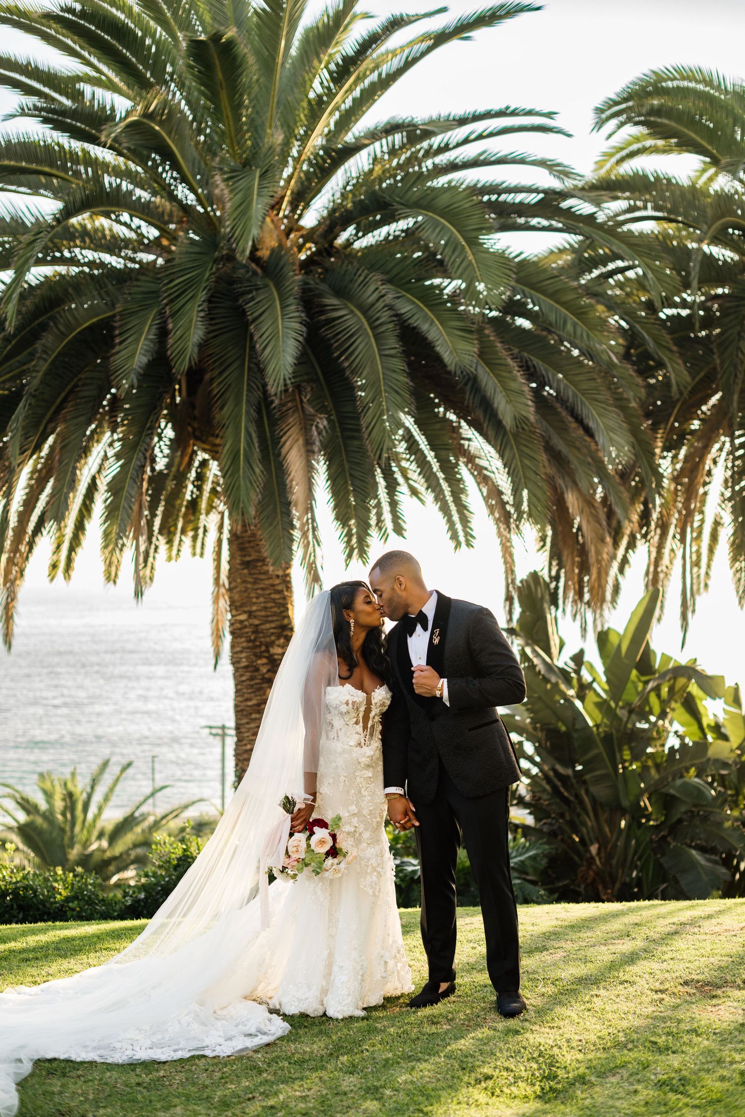 Wedding couple sharing romantic moment under palm trees by oceanside at sunset.