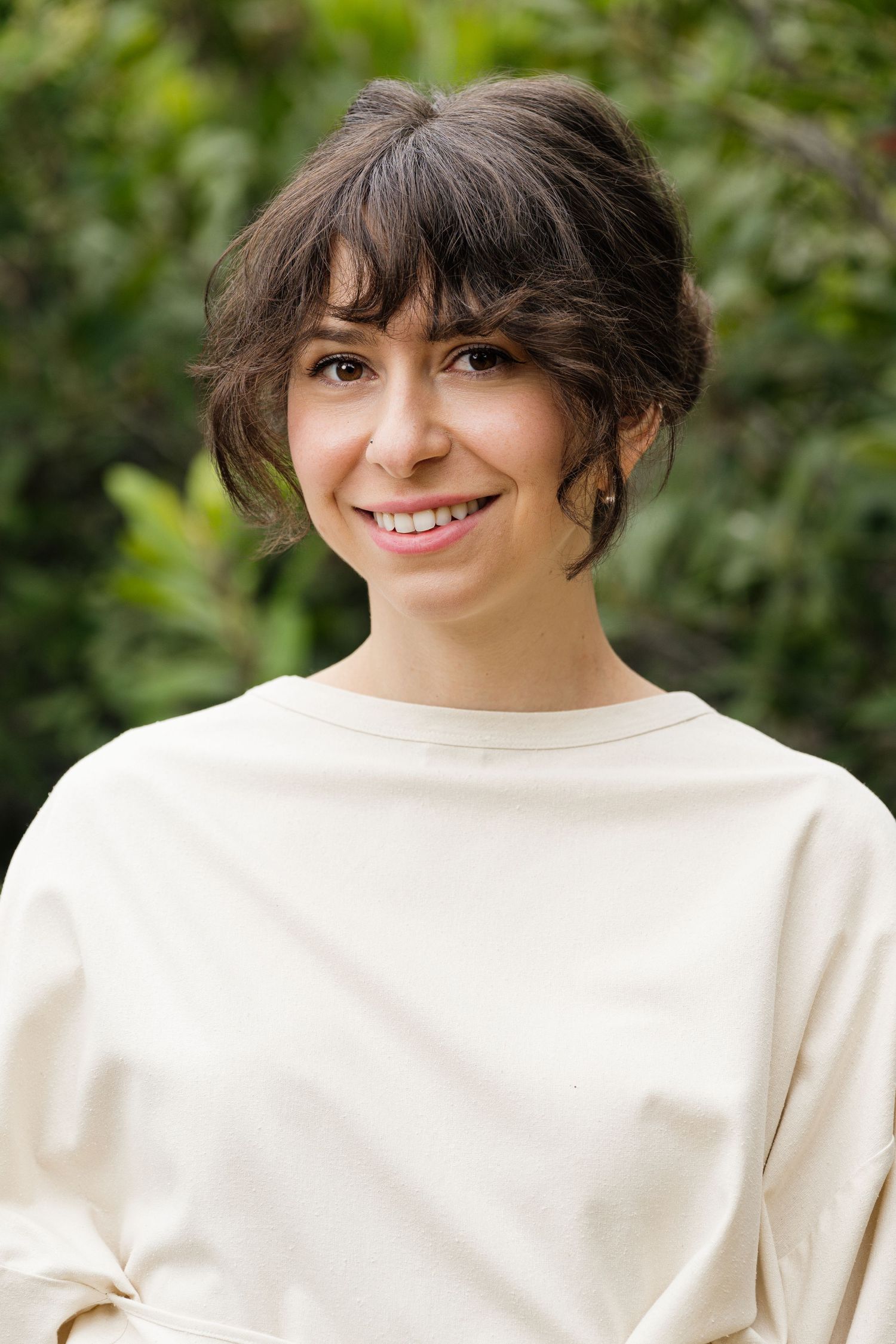 Smiling young woman with short dark hair wearing a cream sweatshirt outdoors with green foliage background.