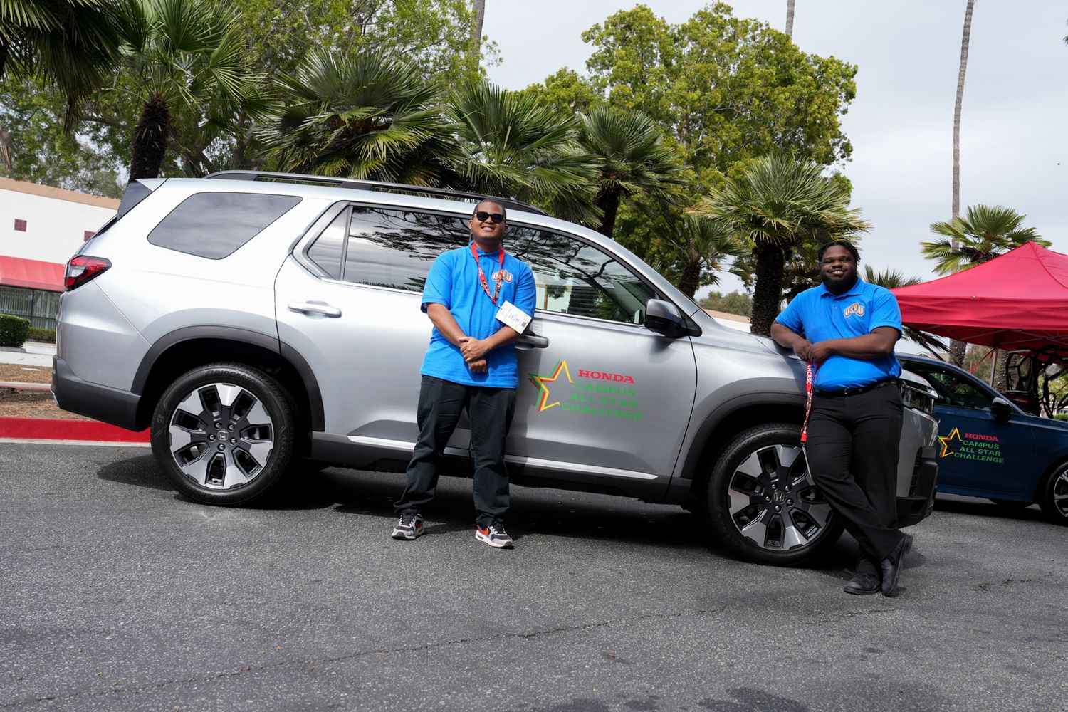 Two men in blue shirts stand beside a silver SUV at an outdoor event on a sunny day.