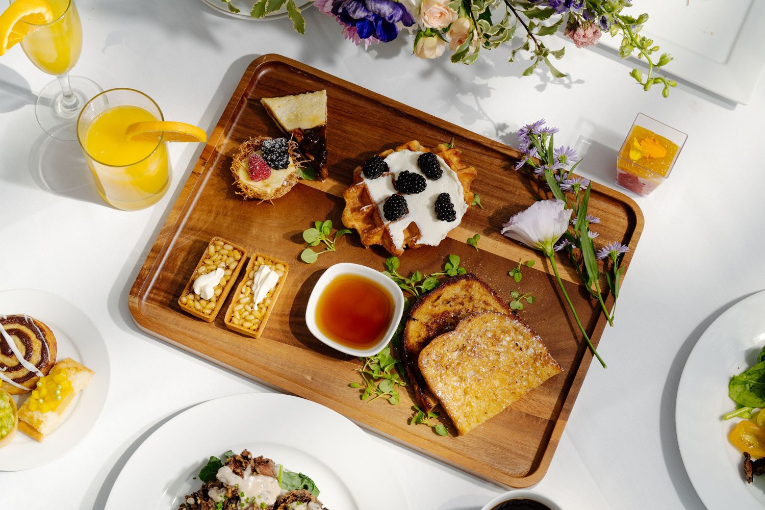 Overhead view of a wooden brunch board with pastries, toast, honey, and fresh flowers on a white table.