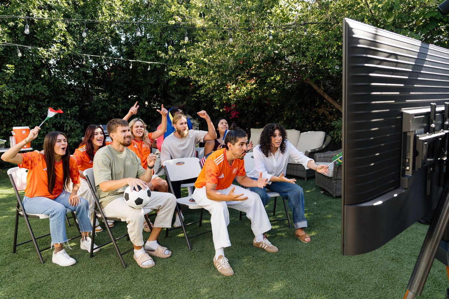 Group of friends in orange shirts cheering while watching sports on outdoor TV in backyard garden.