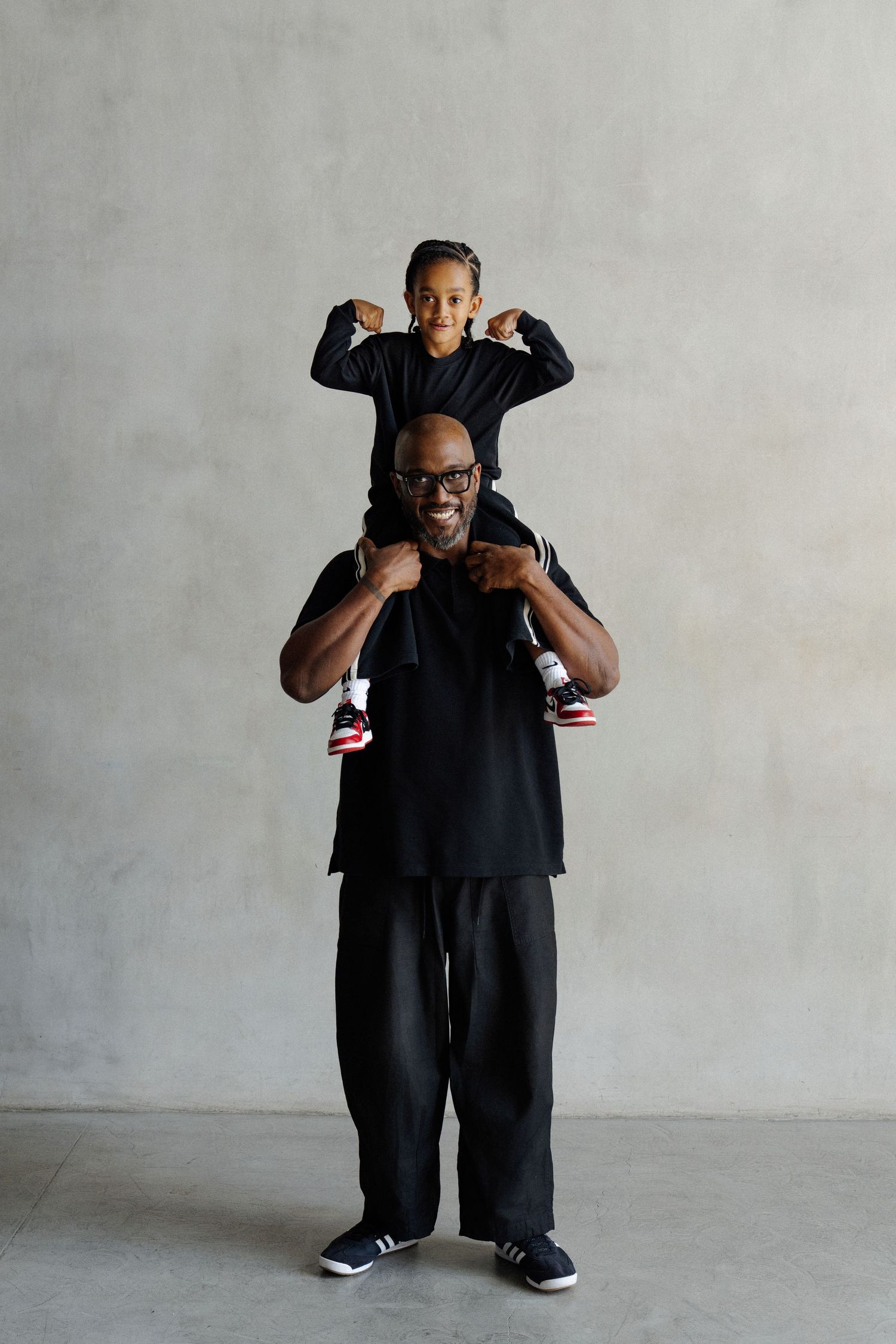 A smiling man carries a young girl on his shoulders, both dressed in black, posing against a gray wall.