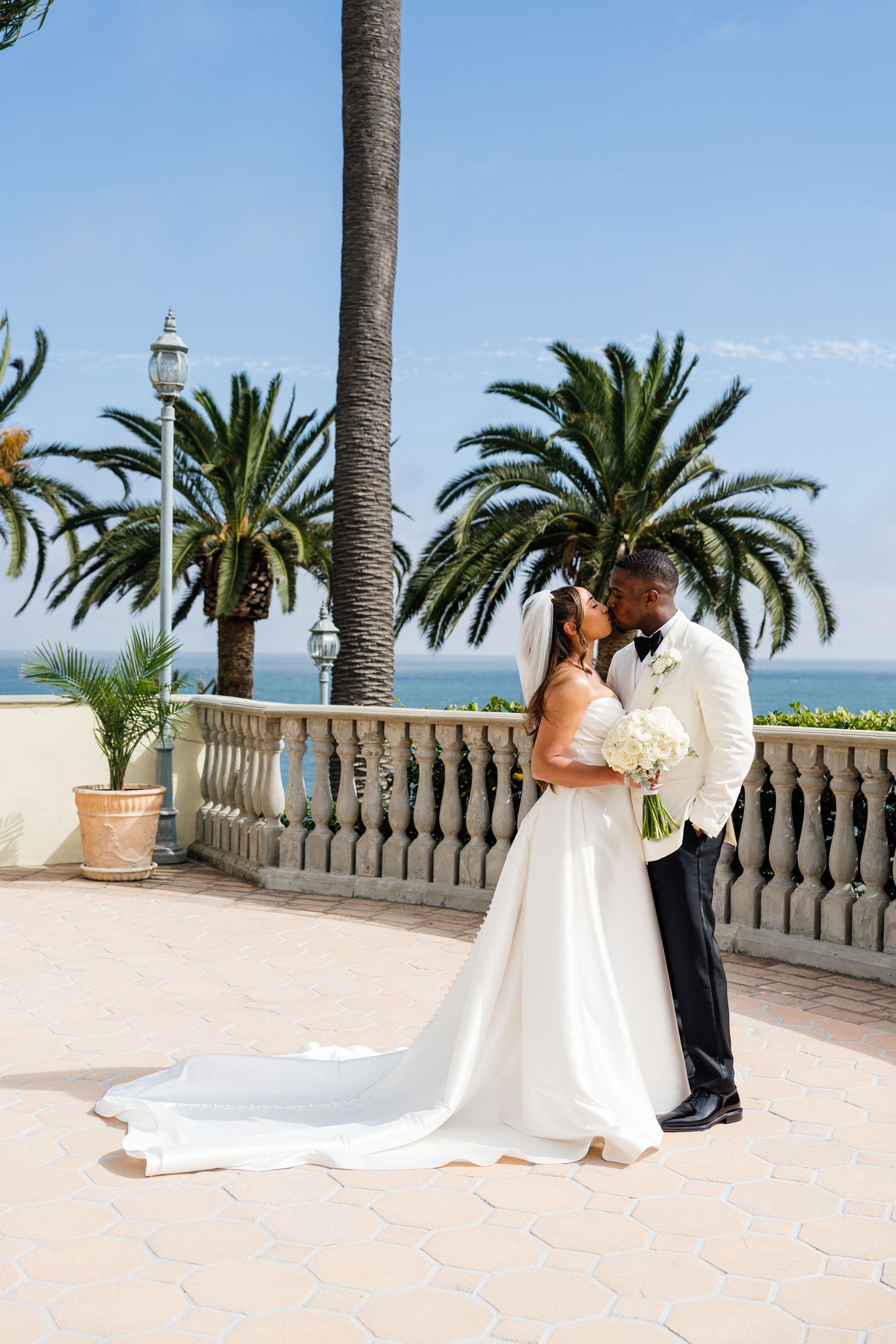 A bride and groom share a romantic kiss on a terrace with palm trees and ocean views on their wedding day.