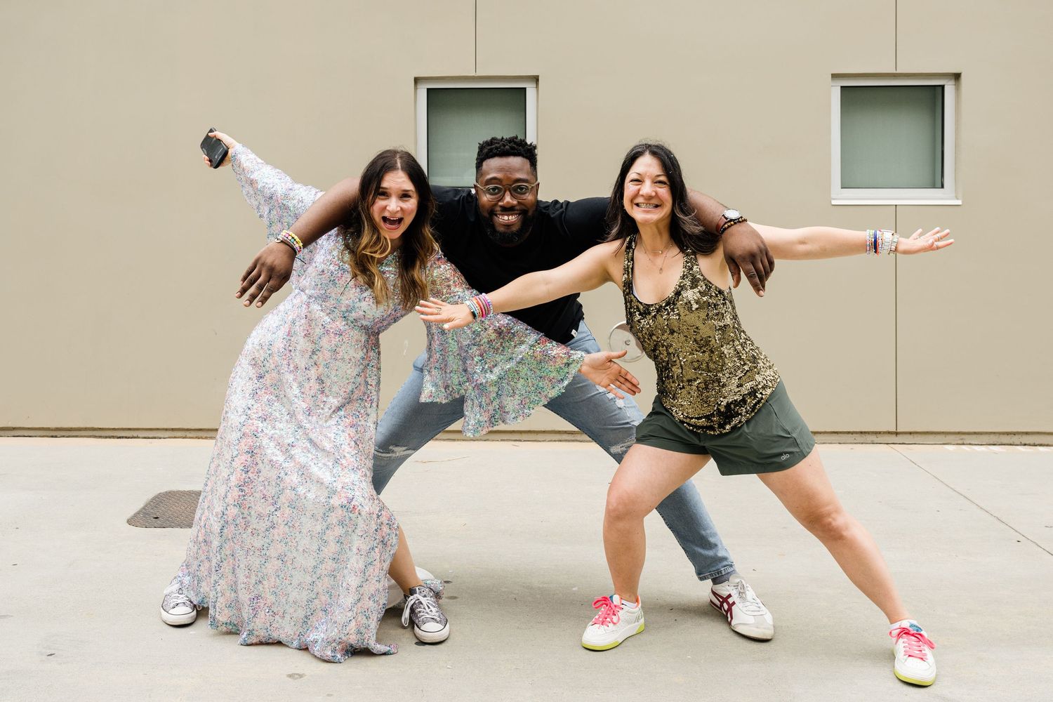 Three people striking playful poses against a beige wall outdoors.