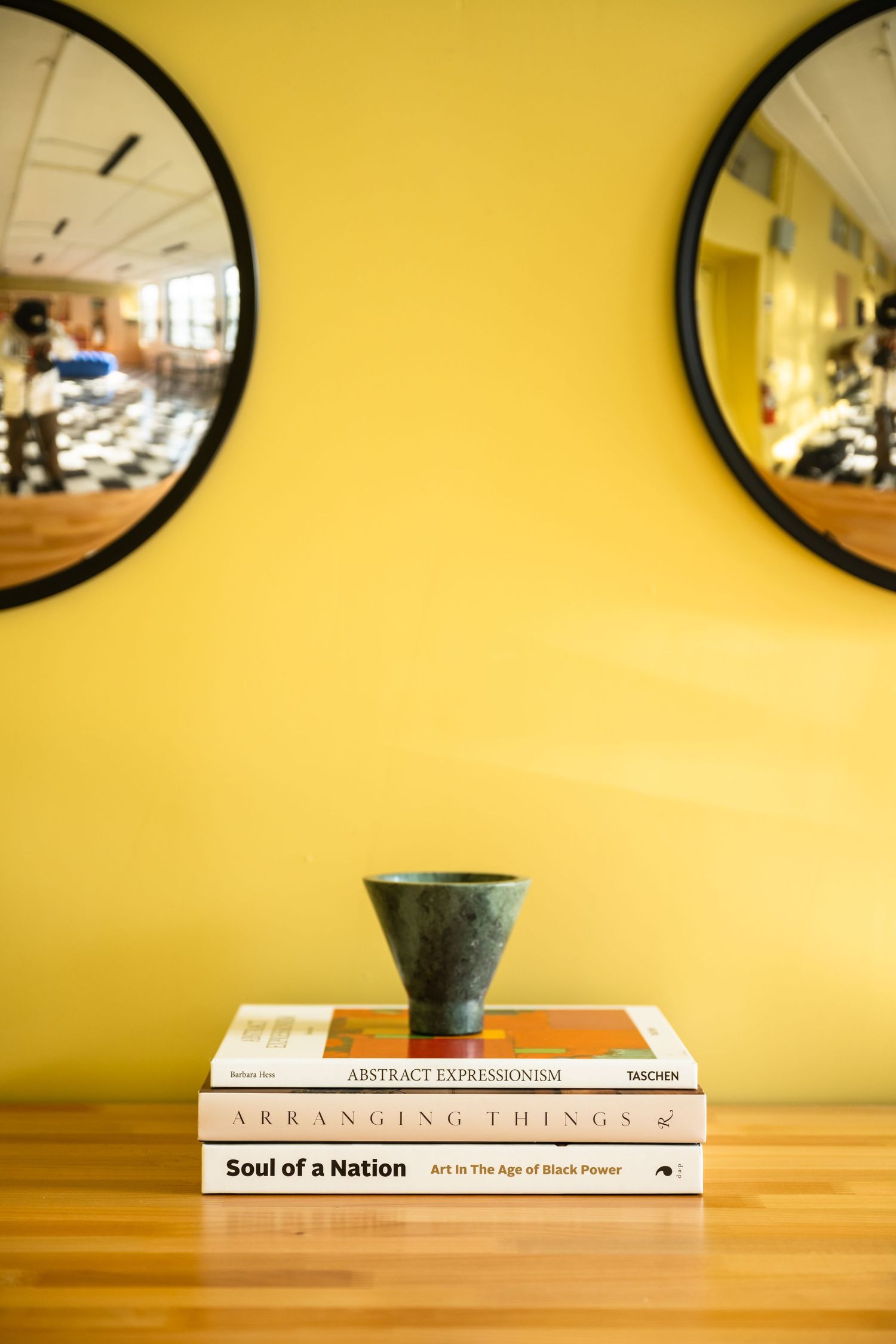 Books arranged on wooden surface with round mirrors on yellow wall above.
