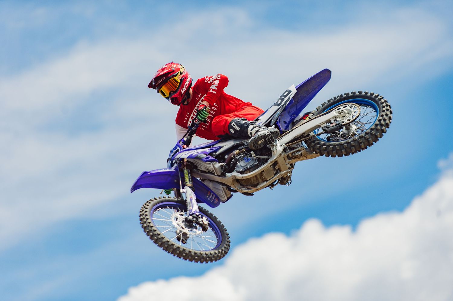 Motocross rider performs aerial stunt on purple dirt bike against blue sky with clouds.