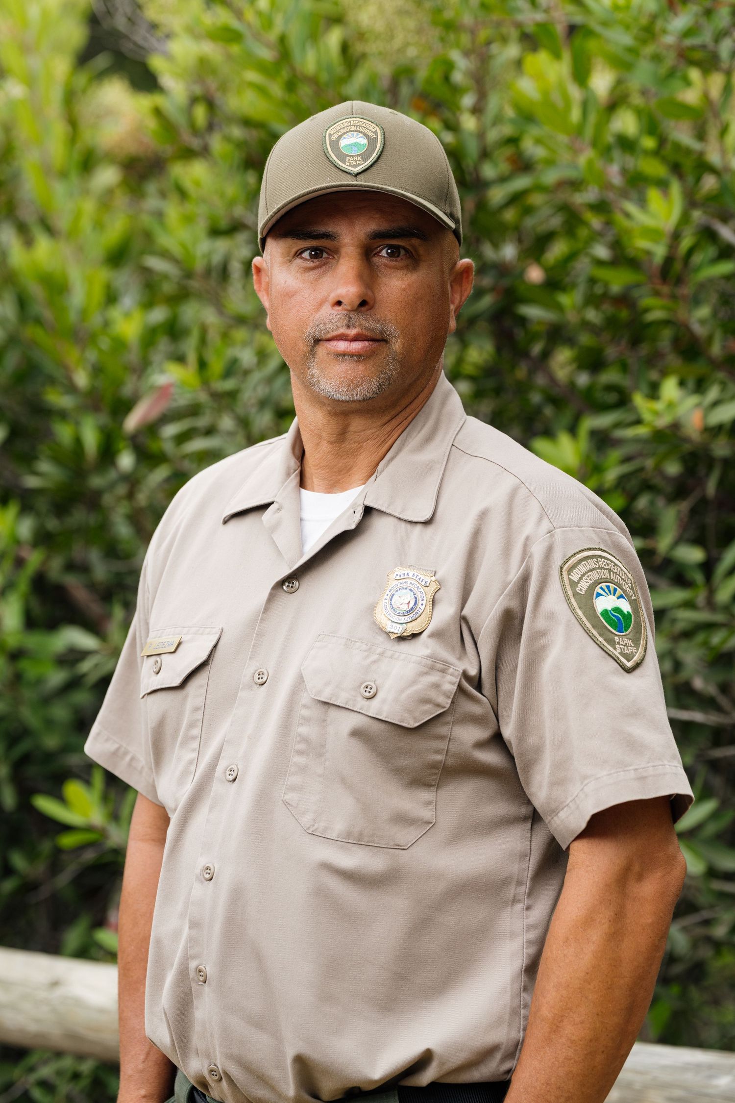 A park ranger in uniform with badge and cap stands outdoors near green foliage.