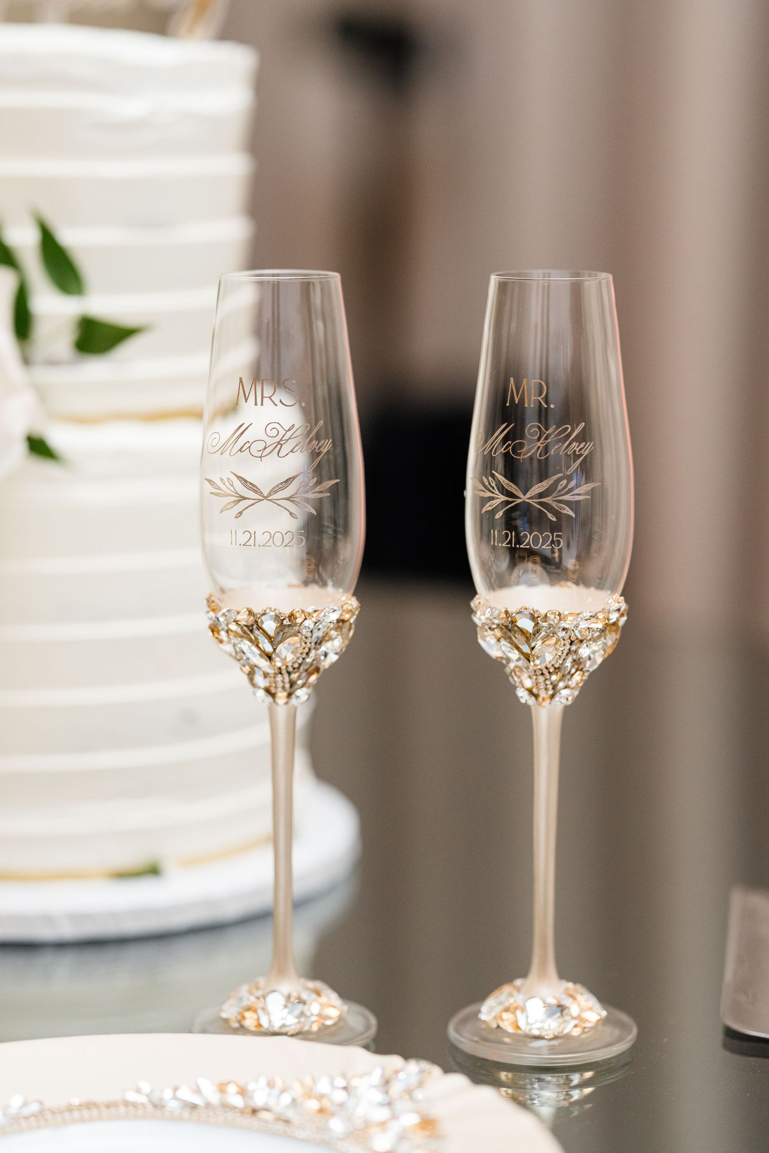 Personalized wedding champagne flutes with gold jeweled stems engraved with Mr. and Mrs. beside a white tiered cake.