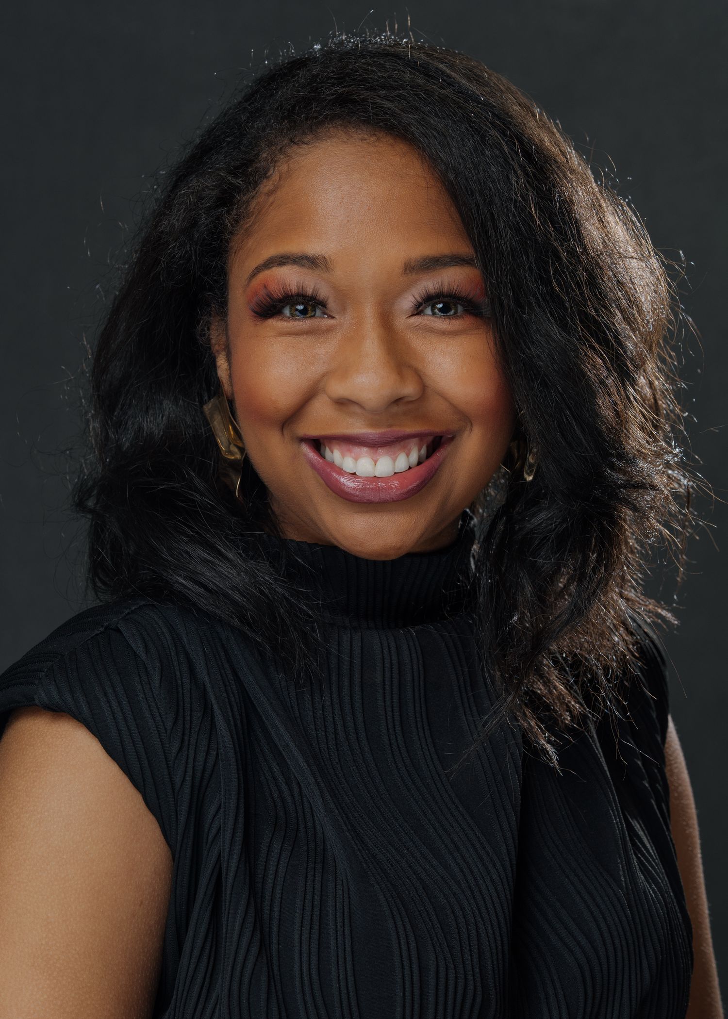 Professional headshot of a smiling woman with long black hair wearing a black turtleneck top against a dark background.