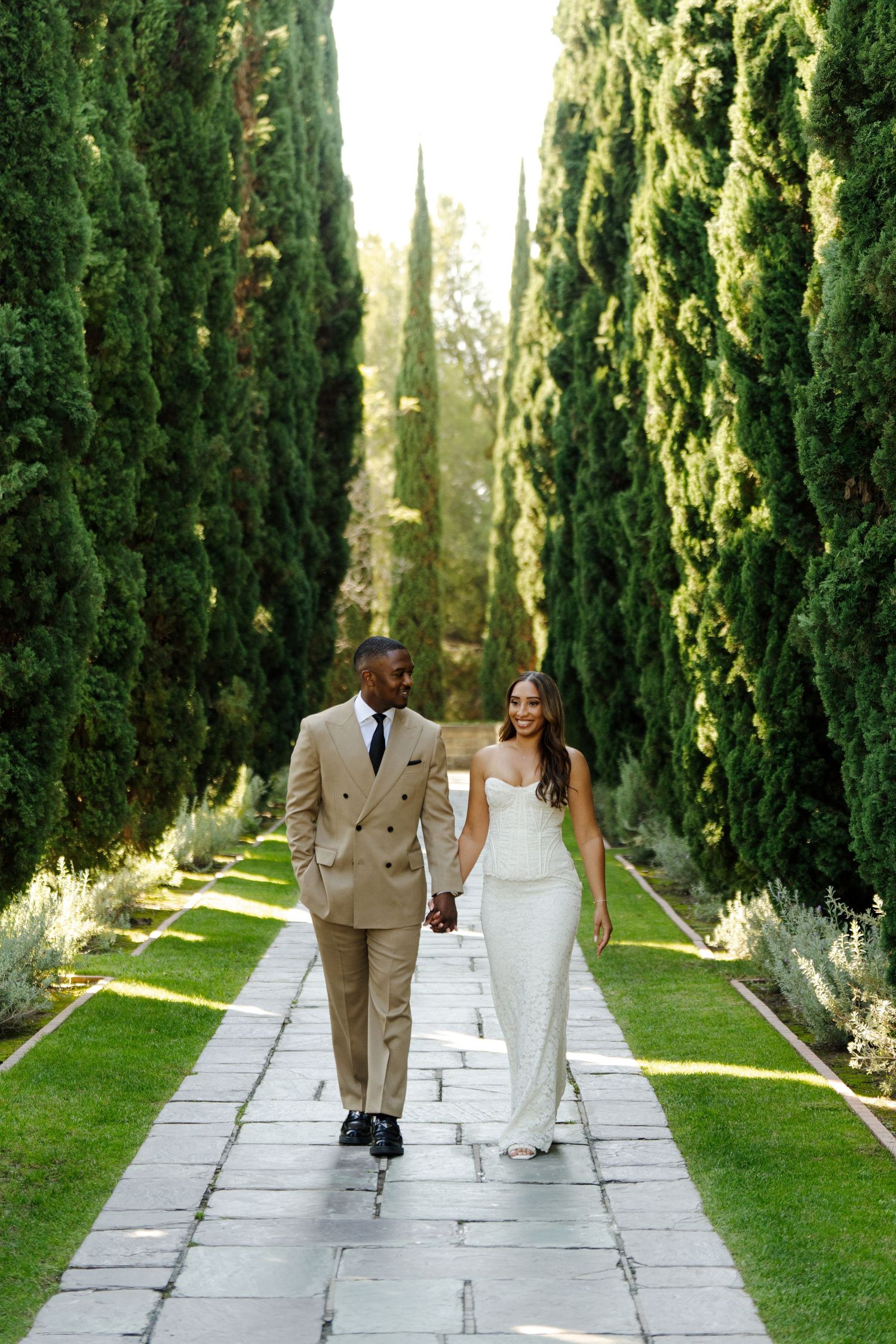 Couple in wedding attire walking down cypress tree-lined path in garden setting.