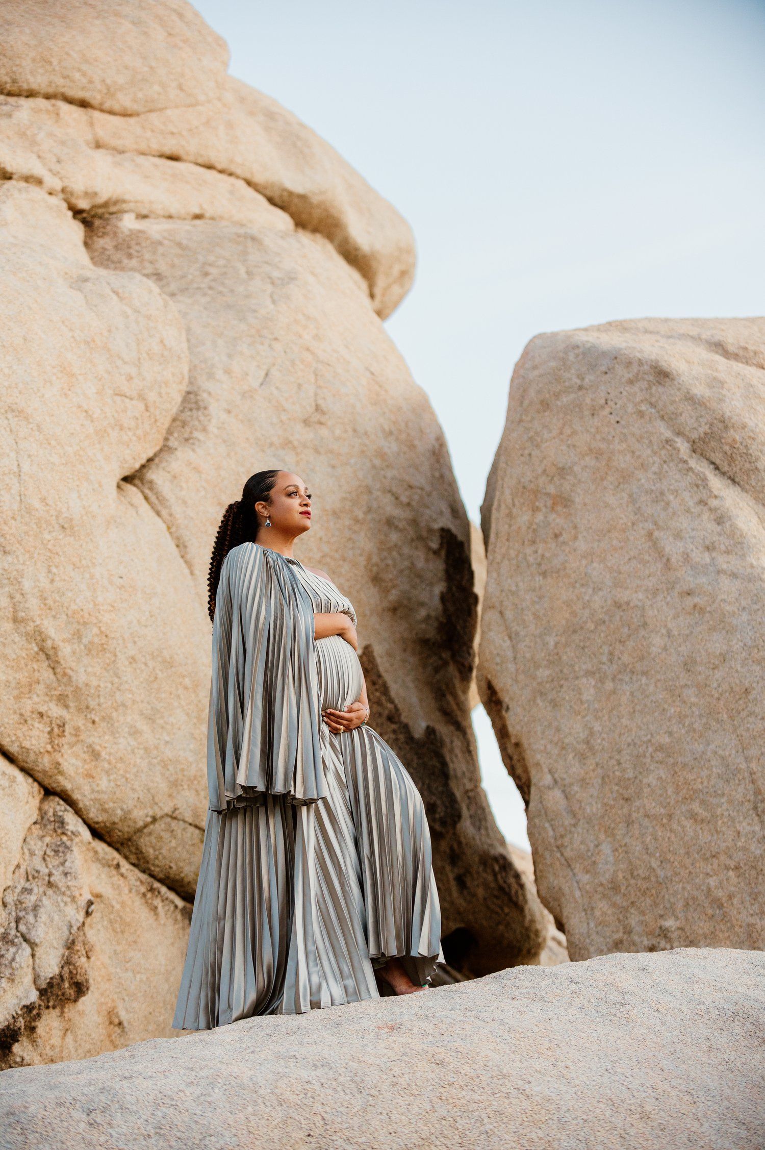 Expectant mother in flowing grey dress poses dramatically among desert rock formations.