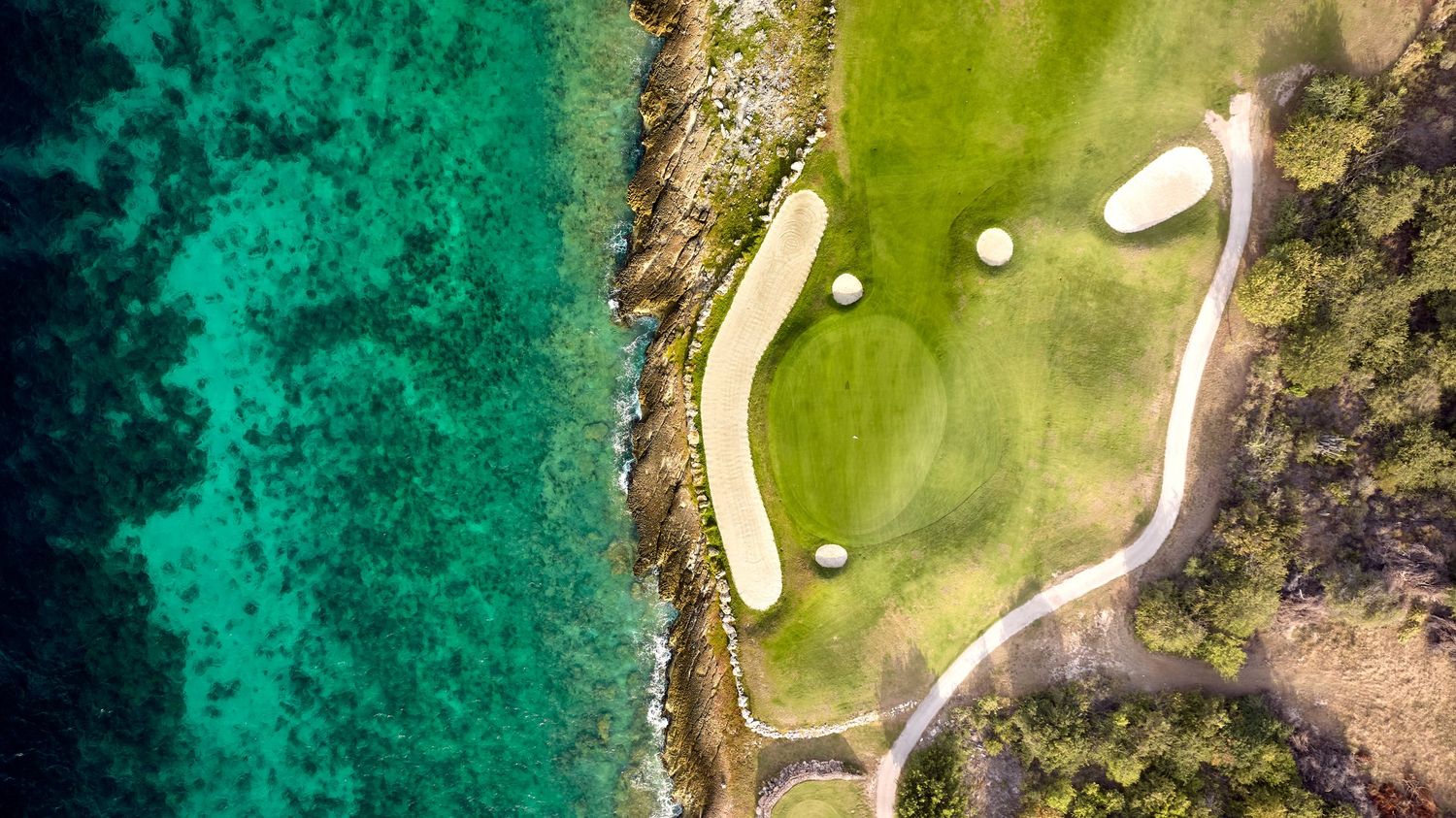 Aerial drone view of a coastal golf course with emerald green fairways bordered by turquoise ocean waters.