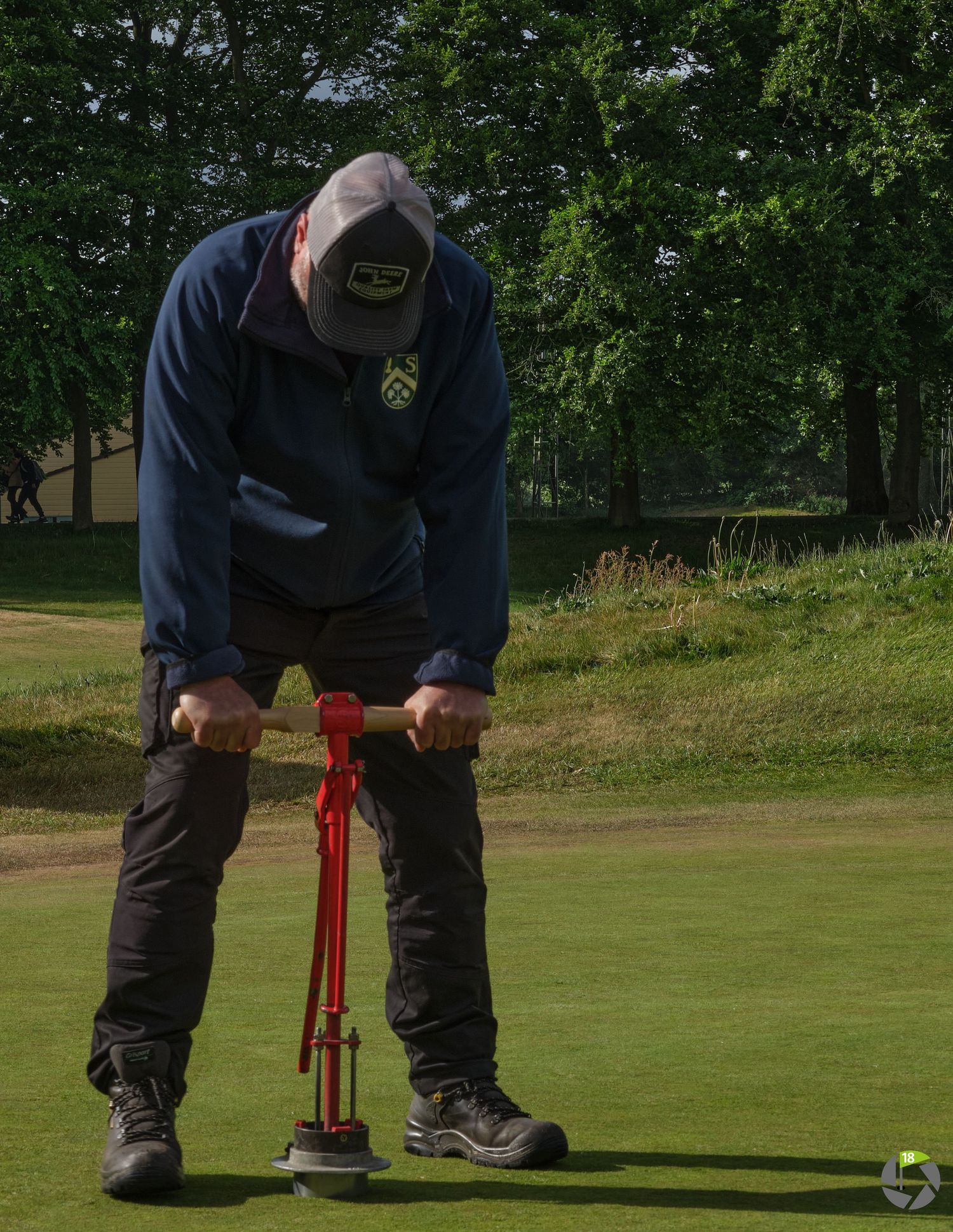 Groundskeeper using a red hole cutter tool on a golf course green, surrounded by trees.